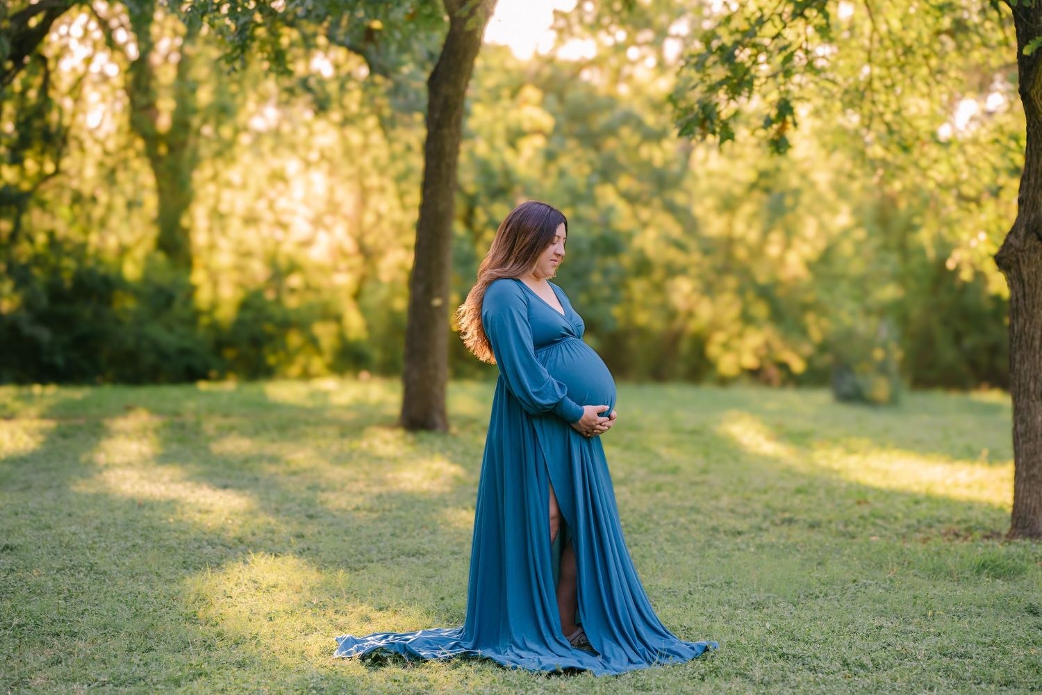 Pregnant woman in teal maternity gown standing in sunlit park, cradling baby bump during outdoor maternity photoshoot.