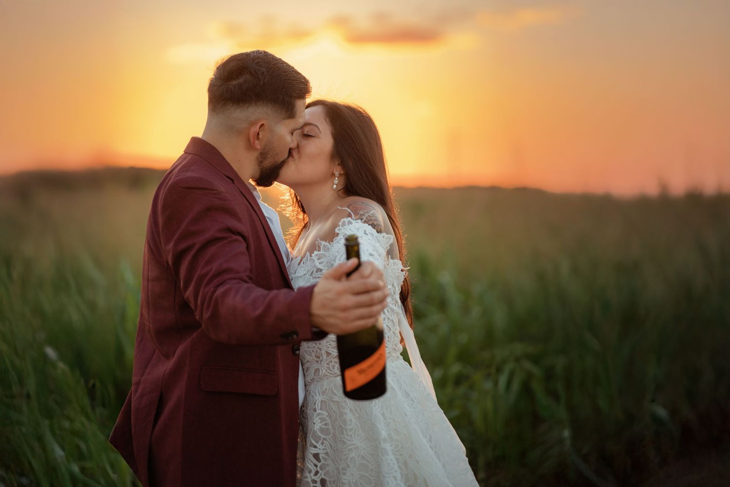 Bride and groom kissing at sunset in a field, holding a champagne bottle, wearing wedding attire.