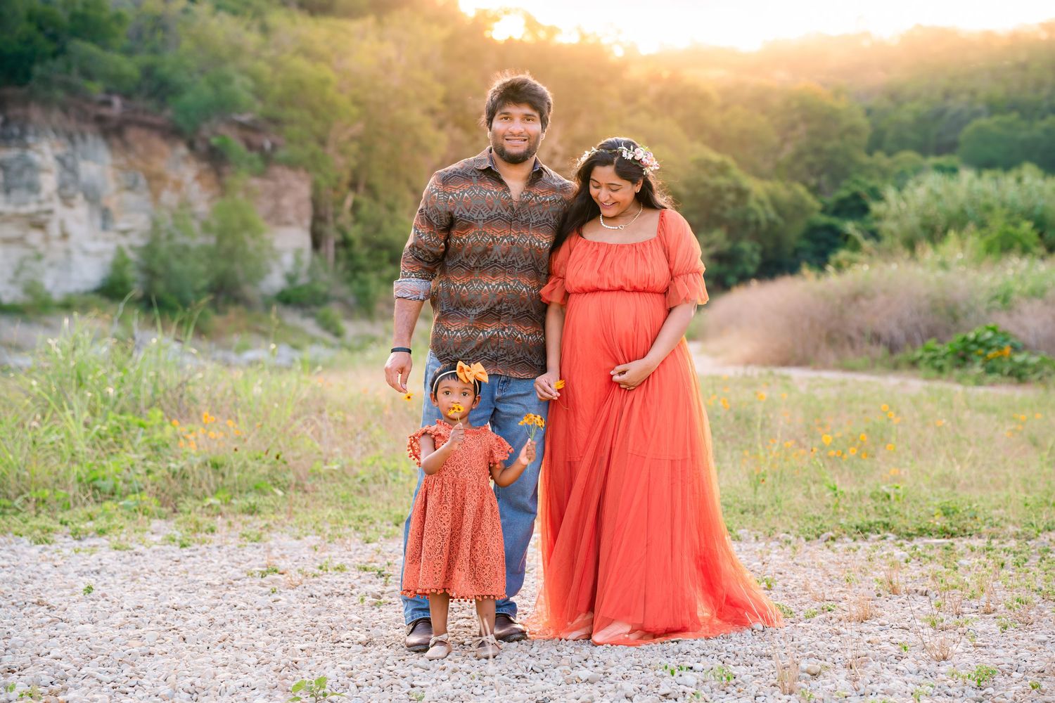Pregnant woman in coral gown with husband and toddler daughter posing outdoors at golden hour maternity photo session.