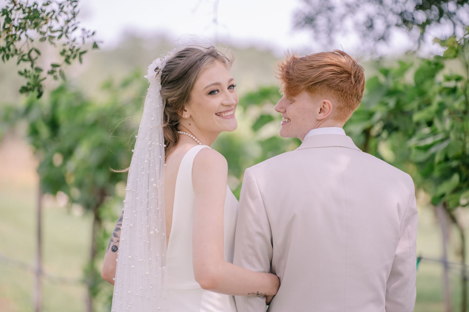 Two people in wedding attire share a tender moment together in a garden setting with greenery and trees in the background.