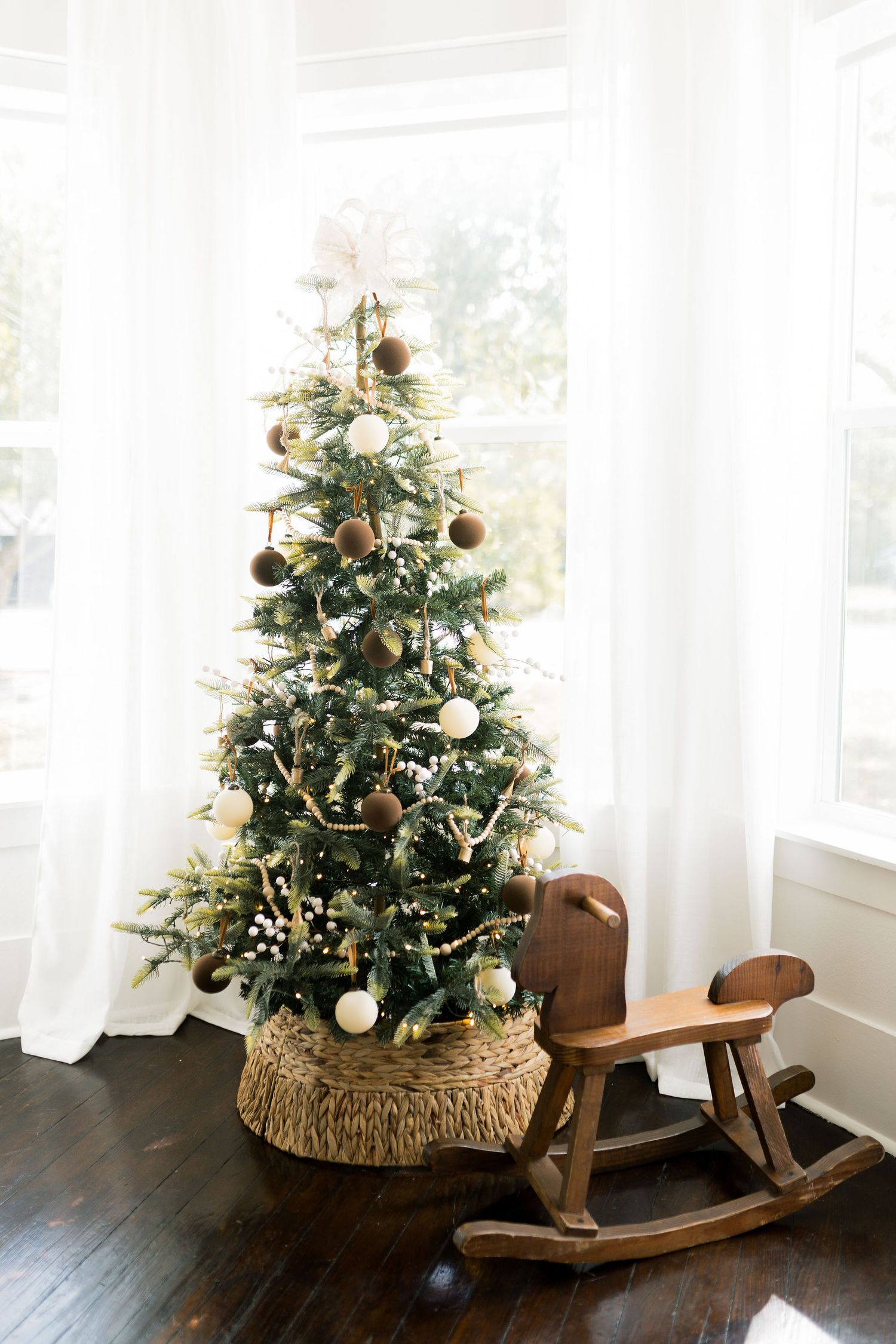 Decorated Christmas tree with gold and white ornaments beside a wooden rocking horse near bright windows.