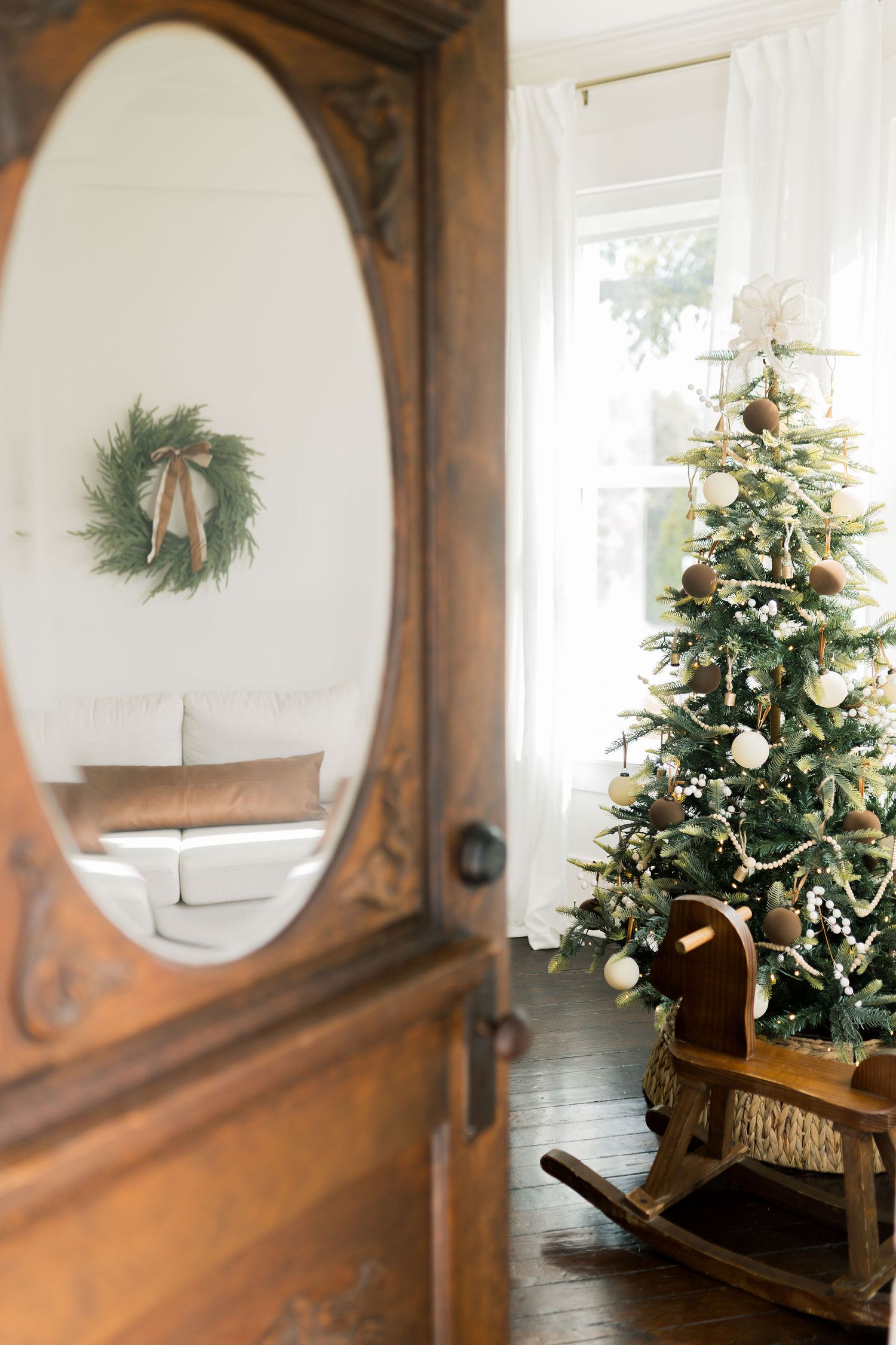 Elegant Christmas living room with decorated tree, wooden rocking horse, antique mirror door, and wreath on white wall.