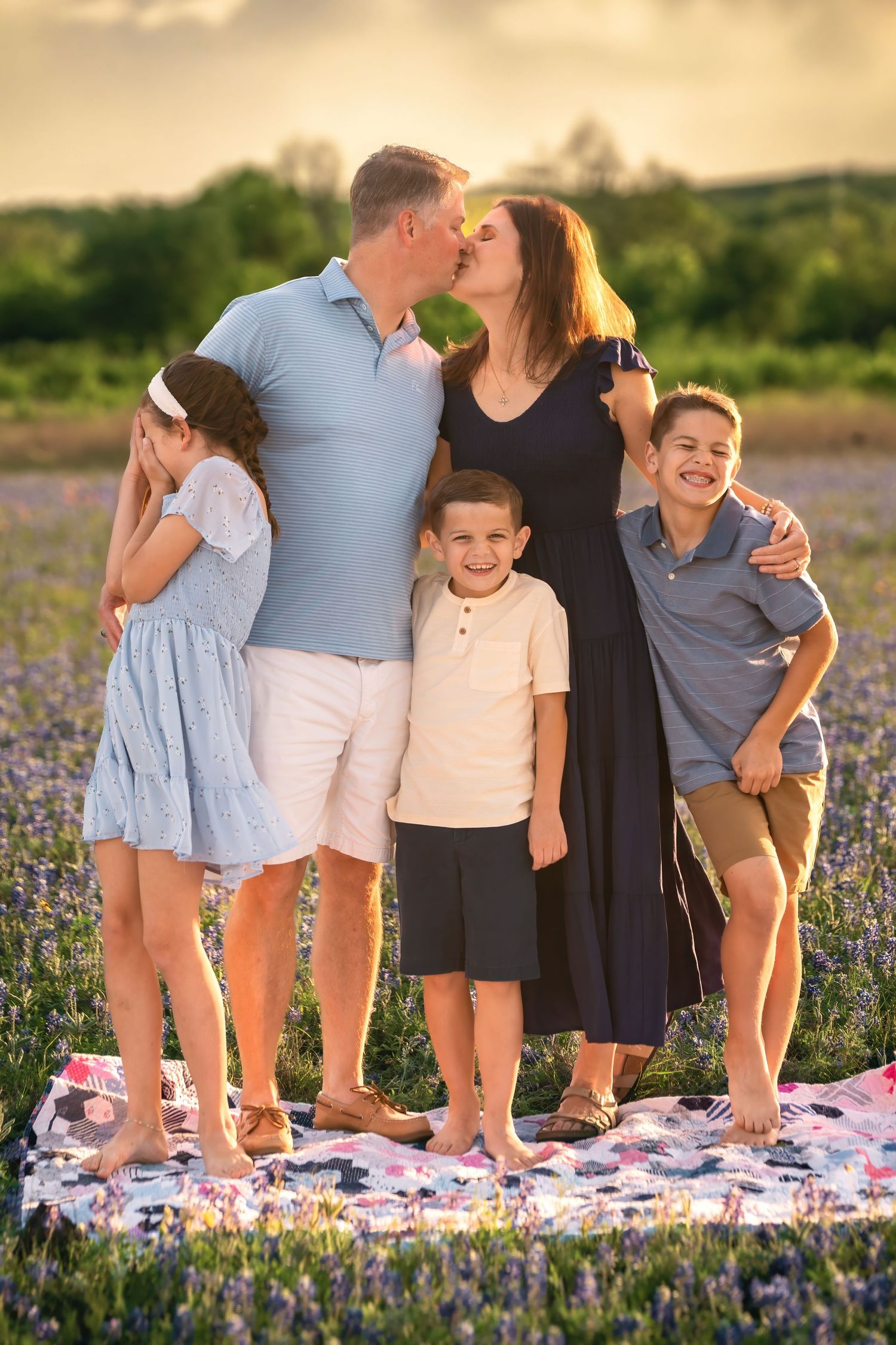 A family shares a tender moment while standing together on a blanket in a field of blooming bluebonnets at sunset.