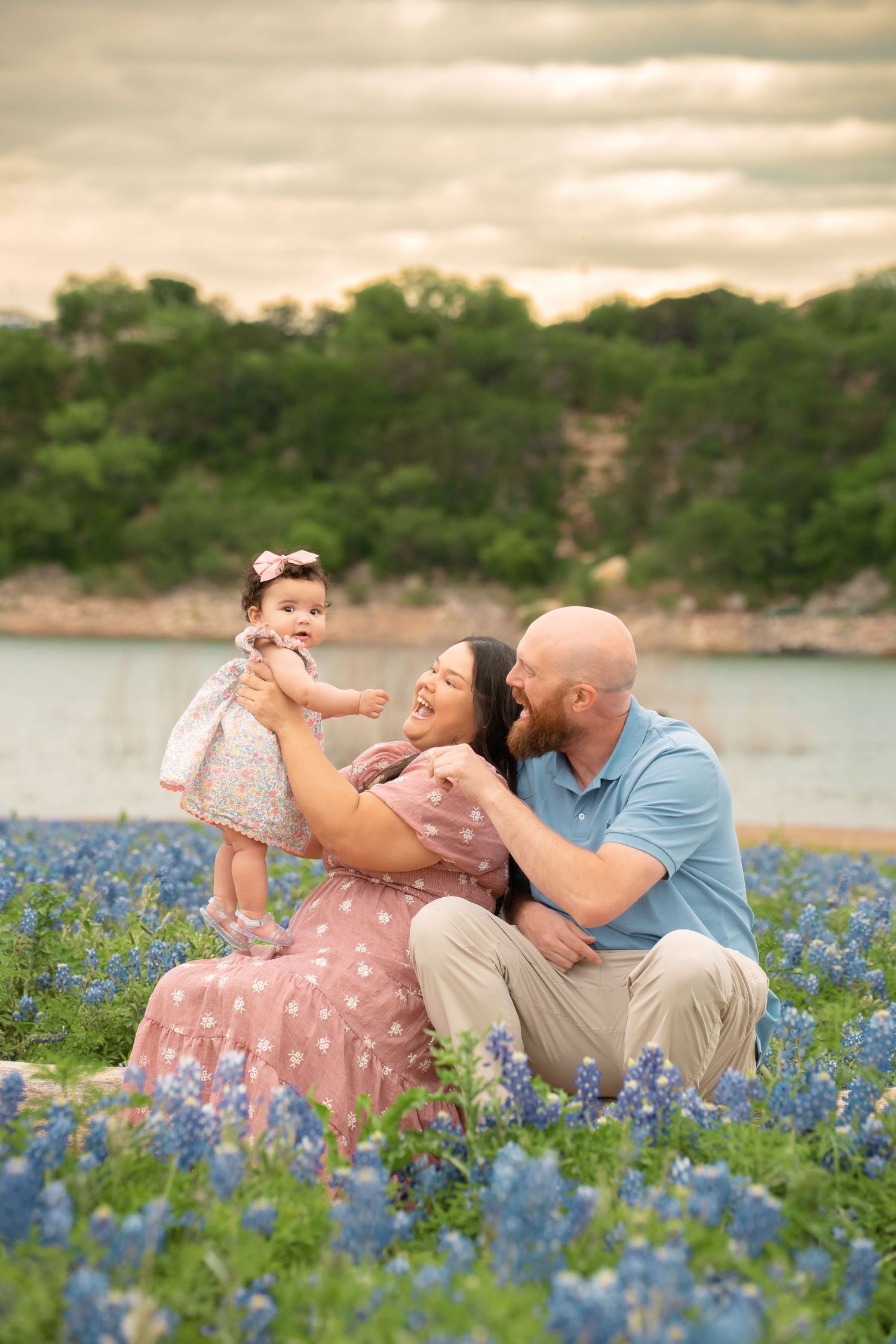 Family portrait in bluebonnet field with parents and baby girl, sitting near lake at golden hour in Texas.