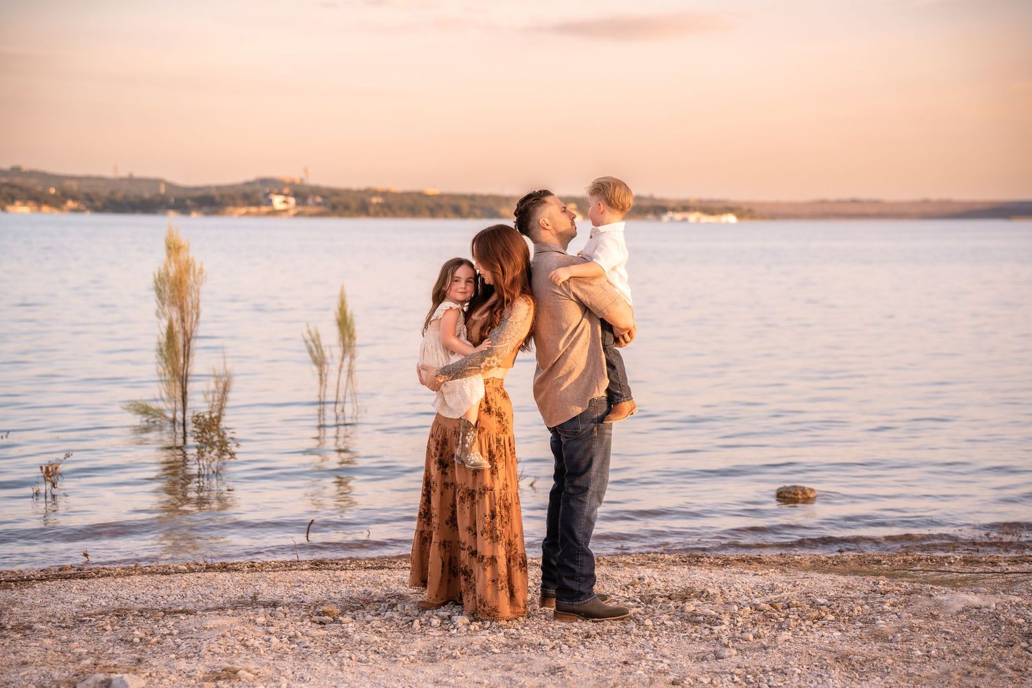 A family shares an intimate moment at sunset along a serene lakeshore with golden light reflecting on the water in Austin, TX