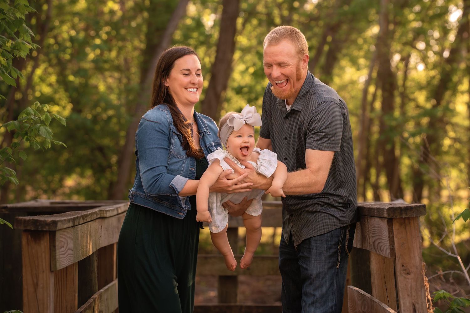 Happy family with laughing baby on wooden bridge surrounded by lush green trees in outdoor portrait.