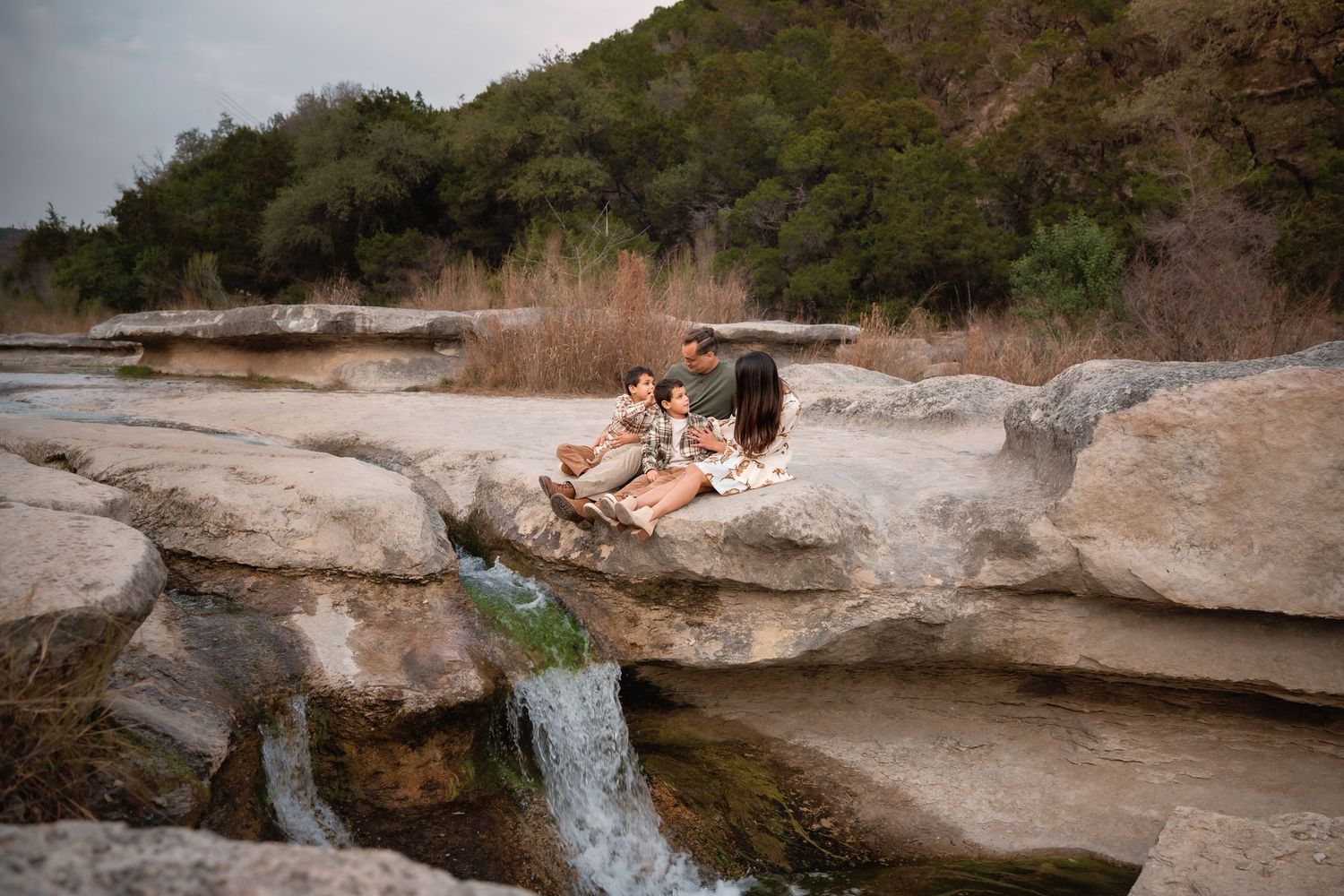 Family sitting on rocky ledge beside a small waterfall surrounded by trees and dry grass in a natural outdoor setting.