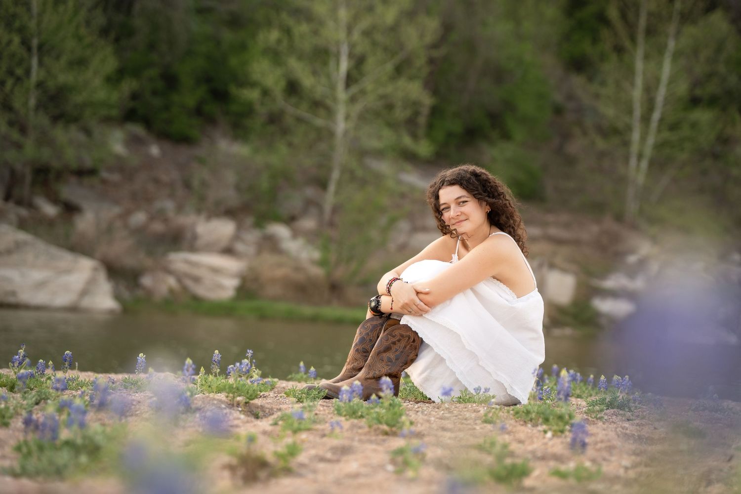 Young woman in white dress and cowboy boots sits among wildflowers by a rocky river.
