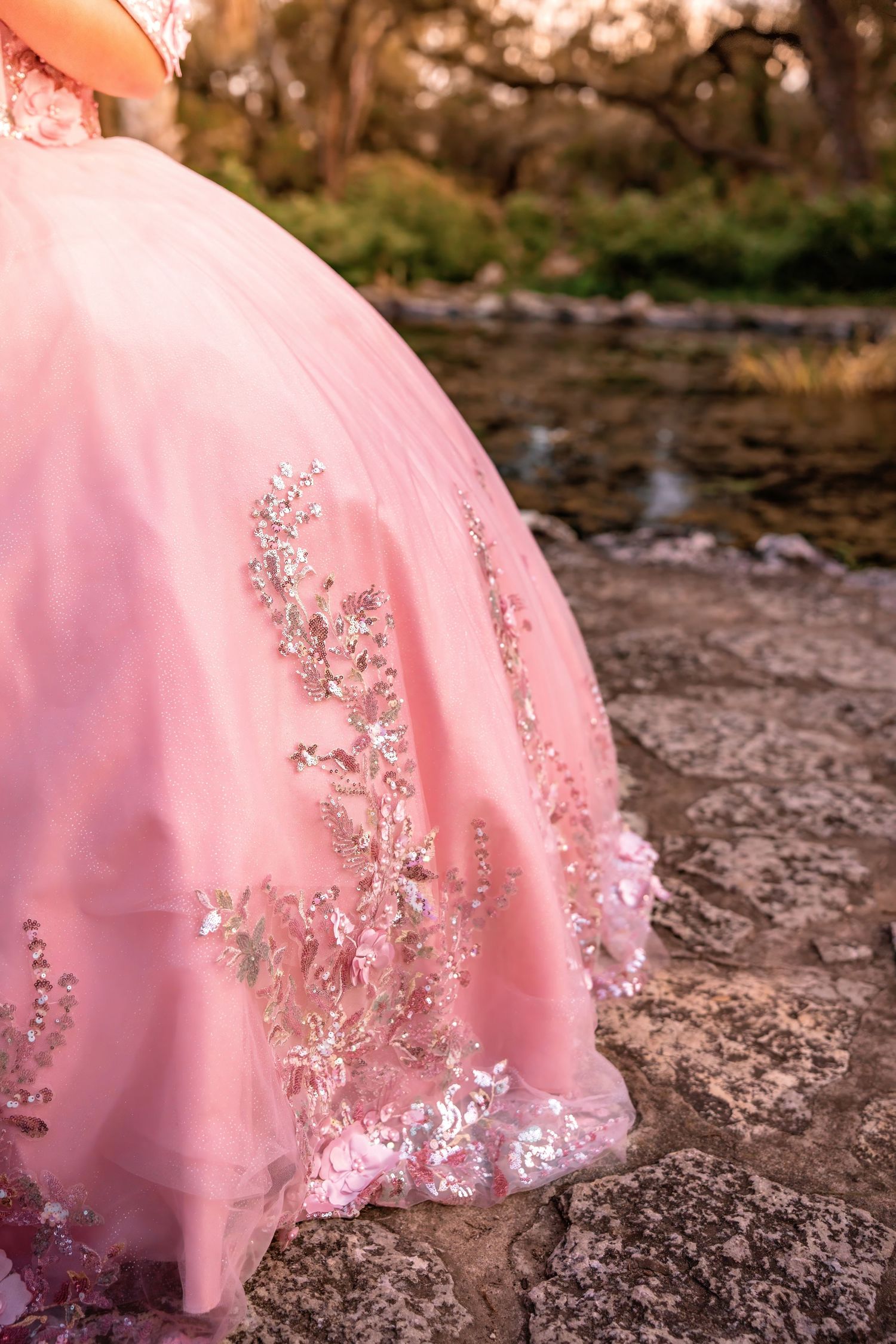 Close-up of an elegant pink quinceañera gown with intricate silver floral embroidery and sequin details on rocky terrain.