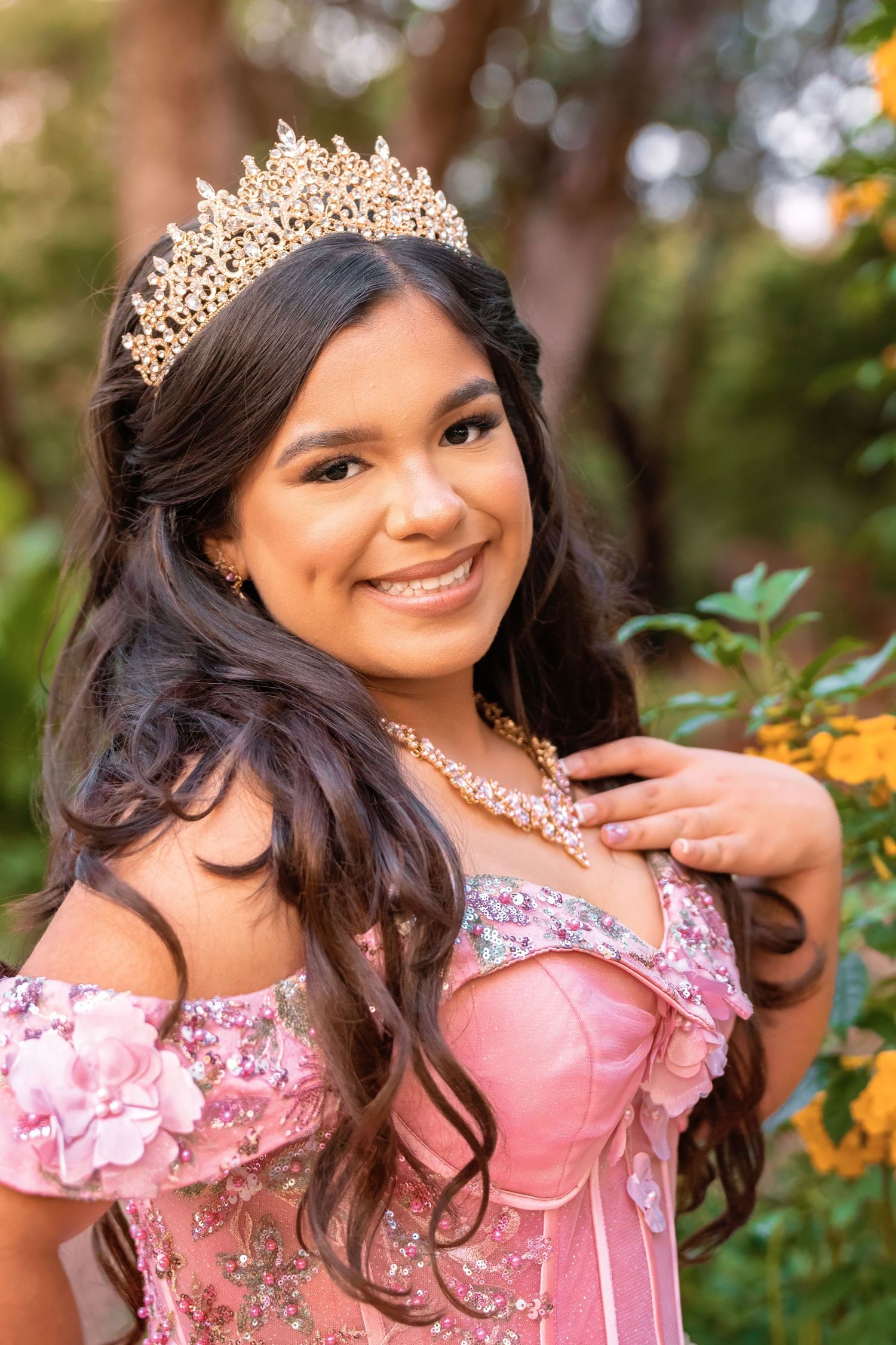 Young Latina girl in pink quinceañera gown and gold tiara smiles outdoors among green foliage.