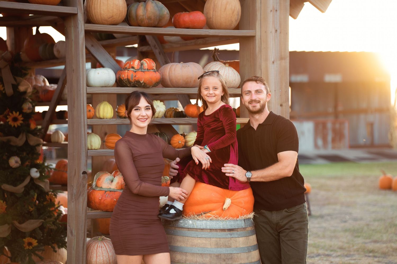 Family posing at a pumpkin patch at sunset, with colorful pumpkins on wooden shelves in the background.