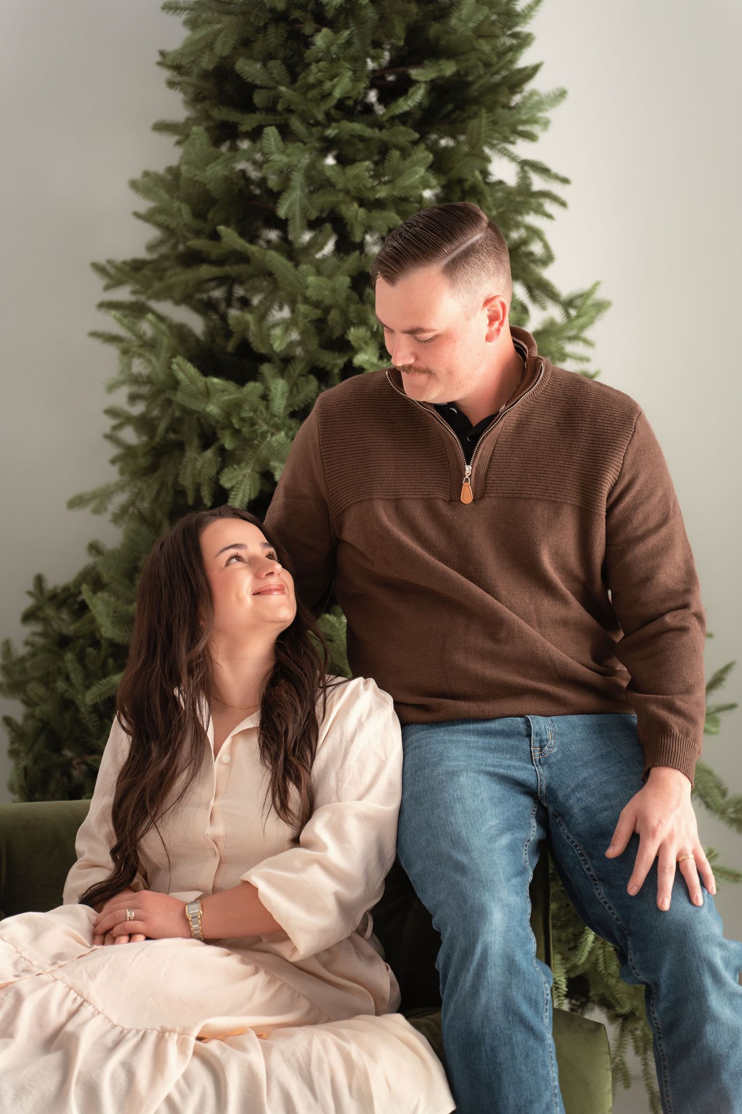 Couple smiling at each other in front of a Christmas tree, woman in white dress, man in brown sweater and jeans.