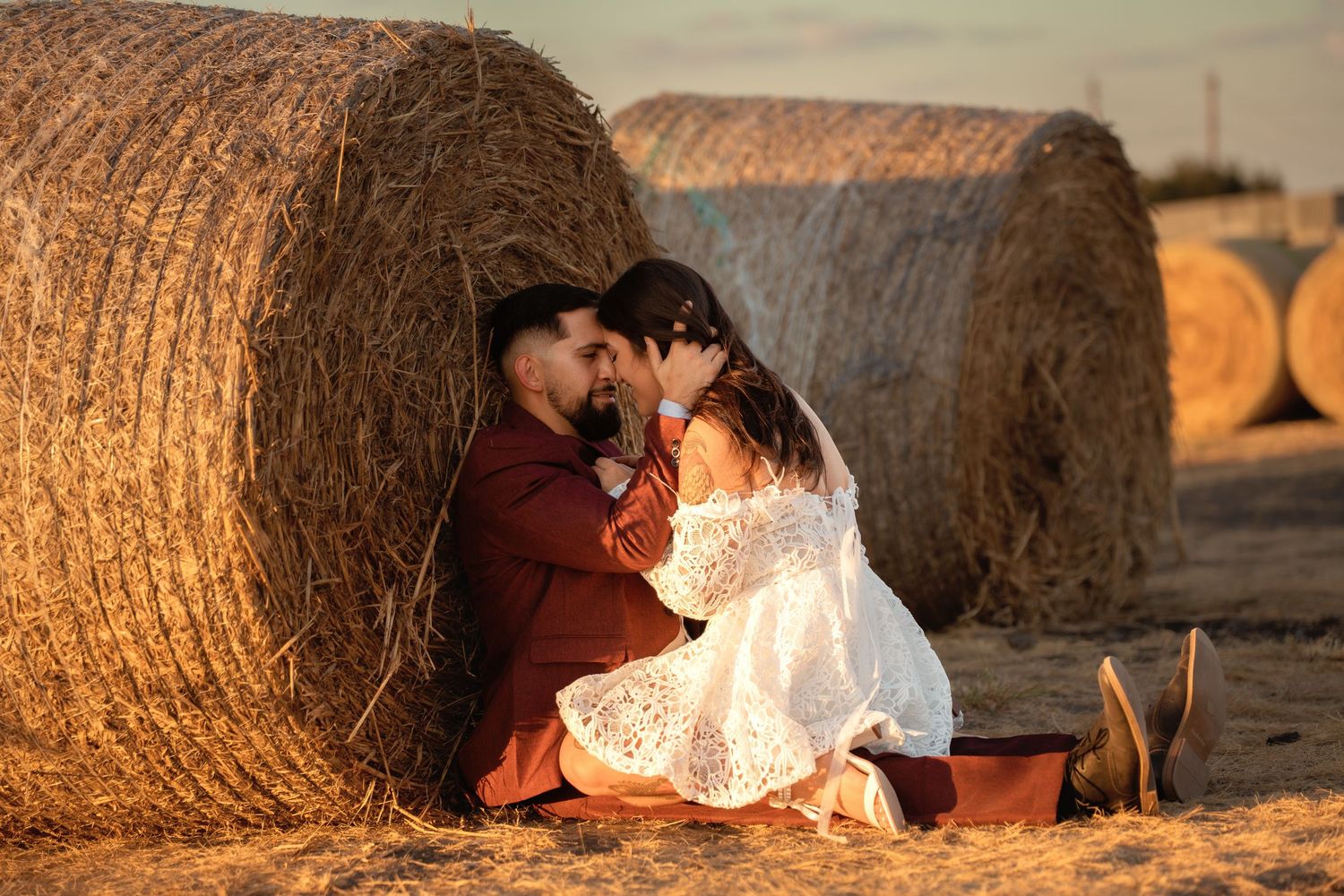 Couple in wedding attire sitting romantically against golden hay bales in a sunlit field at sunset.