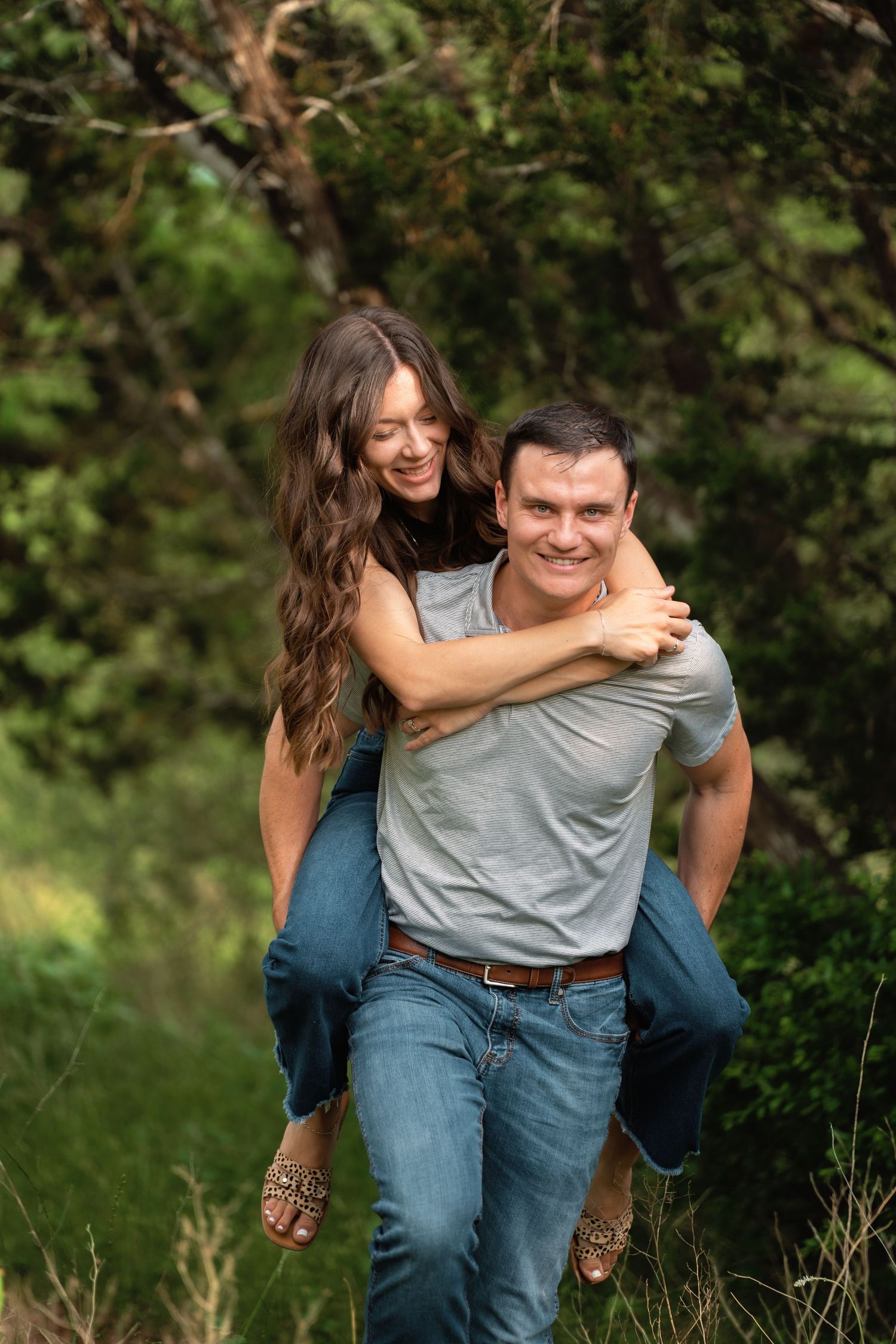 A smiling man gives a happy woman a piggyback ride outdoors surrounded by lush green trees and nature.