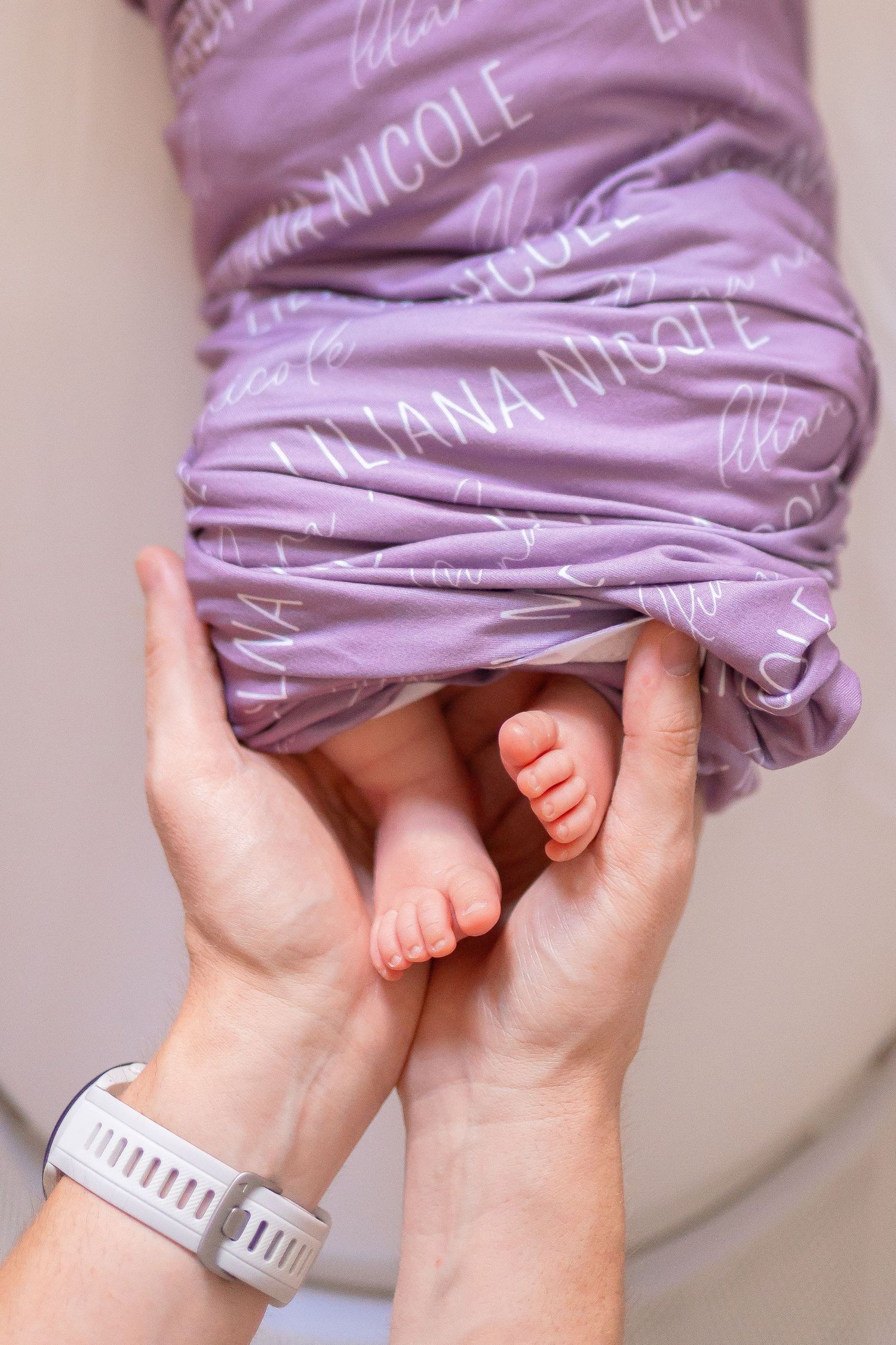Adult hands cradling tiny newborn baby feet wrapped in a purple personalized blanket reading Liliana Nicole.