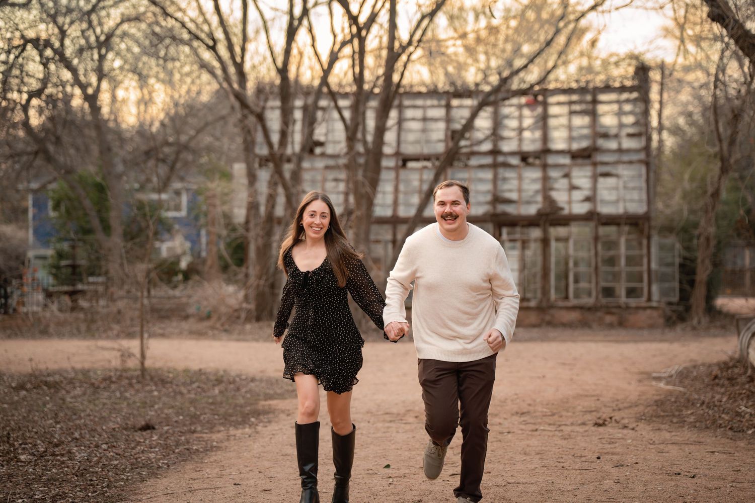 A smiling couple holds hands and runs together outdoors near a rustic building surrounded by bare trees.