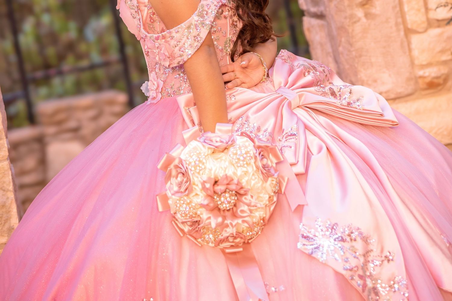 Pink quinceañera ball gown with floral bouquet and large bow detail, worn outdoors near stone wall.