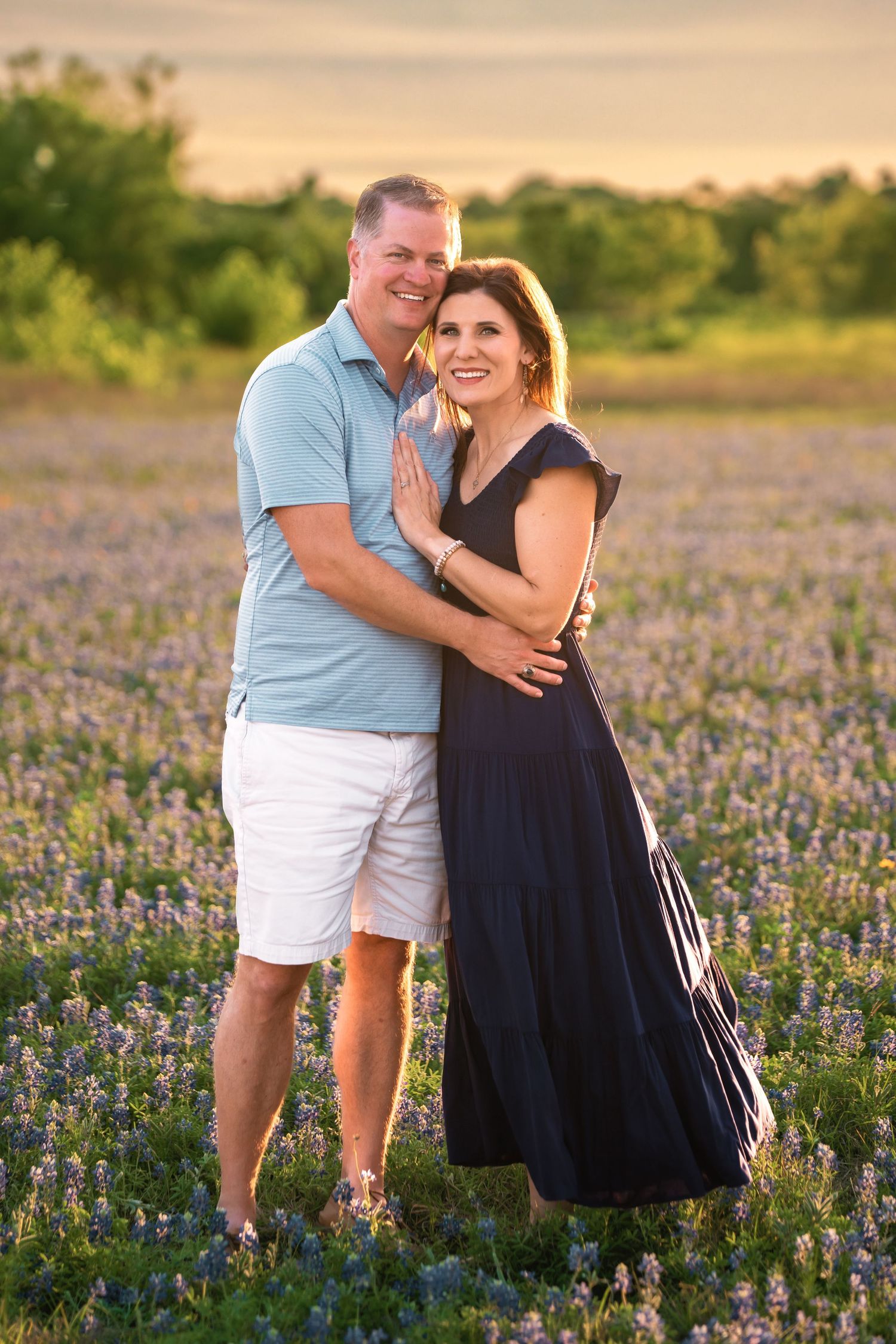 A couple embraces while standing in a beautiful field of purple wildflowers during golden hour sunset.