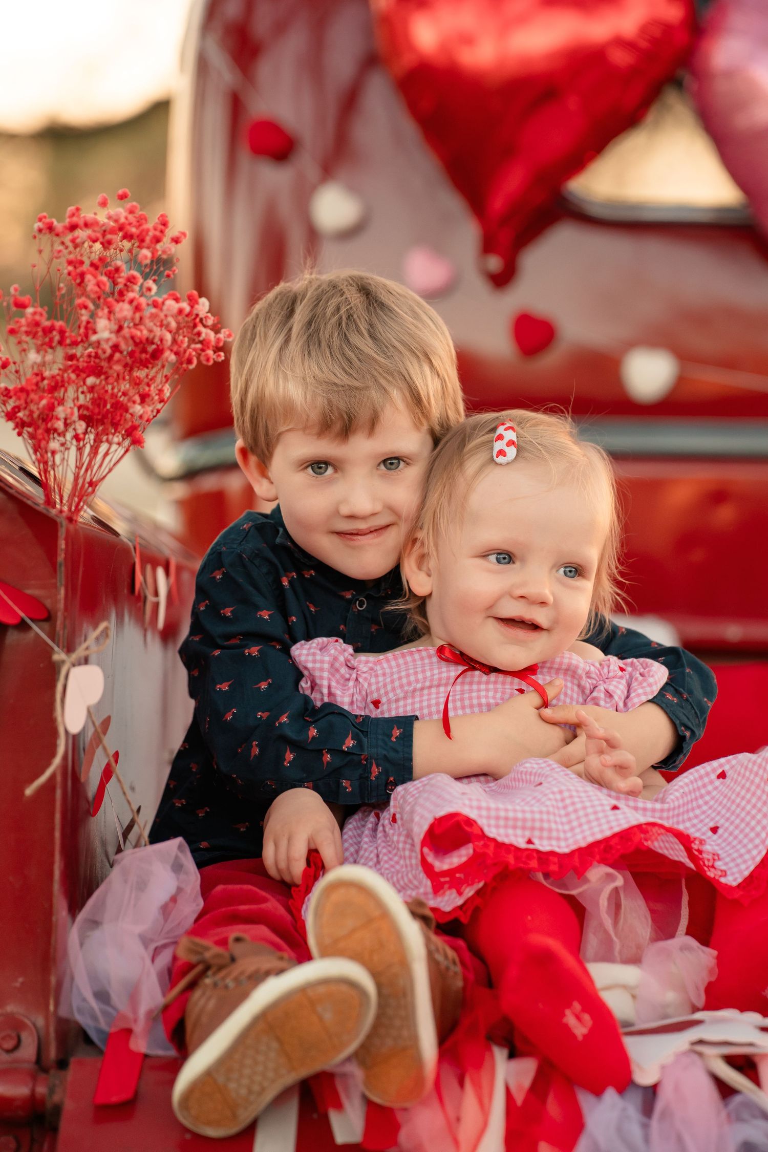 Two smiling children in Valentine's Day outfits sit together on a red vintage truck with heart balloons and red flowers.