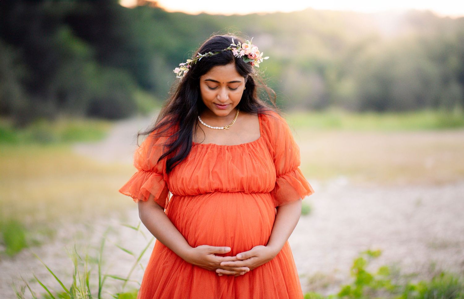 Pregnant woman in orange dress and floral crown cradling baby bump outdoors in soft natural light.