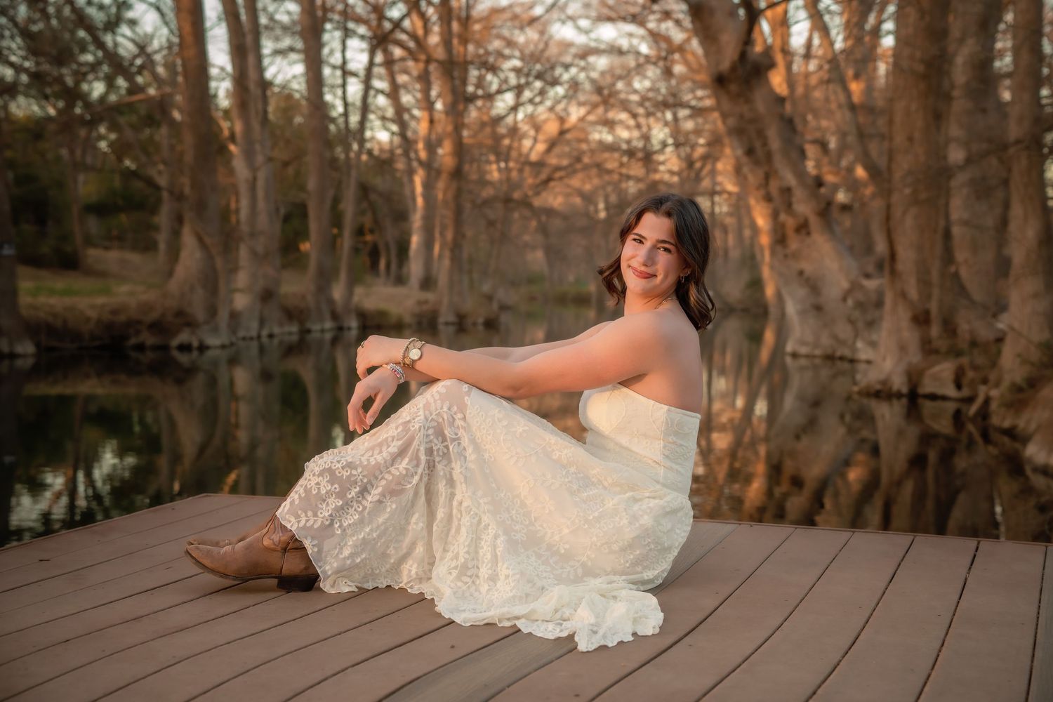 Young woman in white lace dress and cowboy boots sitting on a wooden dock by a reflective pond at sunset.