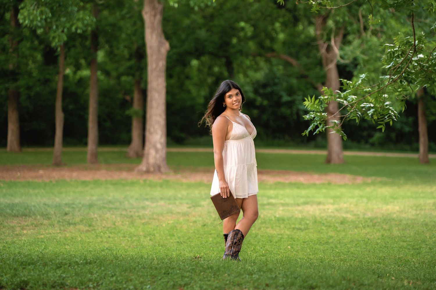 Young woman in white dress and boots holding a purse, smiling in a lush green park surrounded by trees.