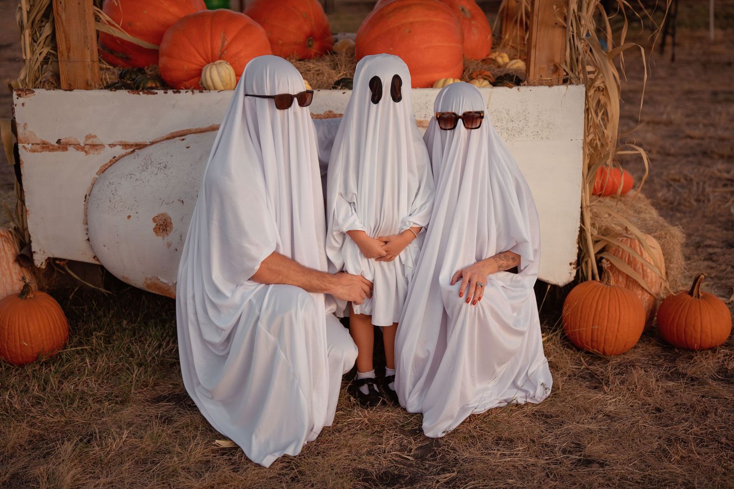 Three people in white ghost costumes with sunglasses pose among pumpkins for Halloween.