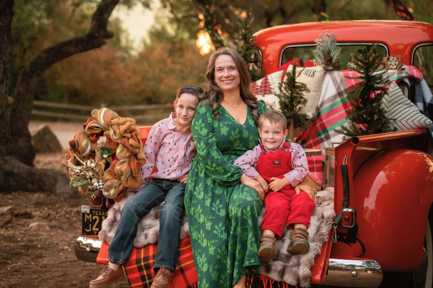 A woman in a green floral dress poses with two children on a decorated red vintage truck for a Christmas photo.