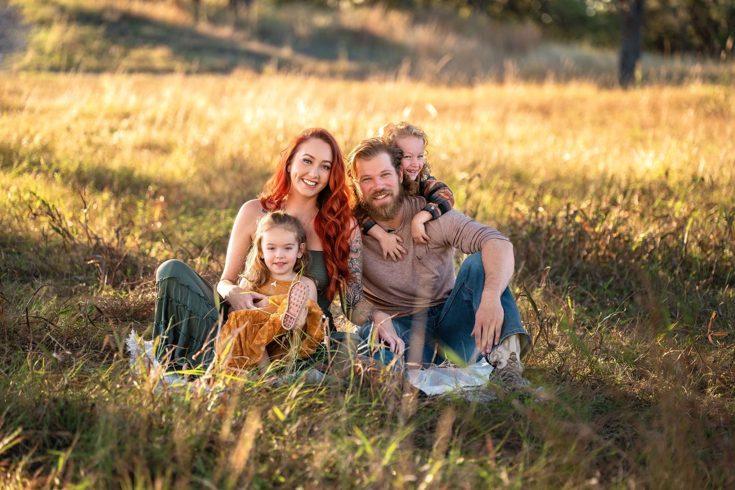 Happy family of four sitting in golden sunlit meadow, parents with red hair smiling with two young children outdoors.