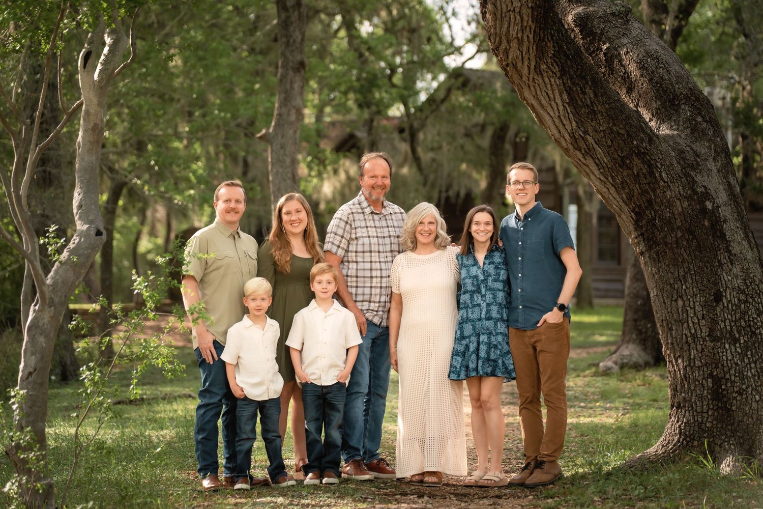 Family of eight poses outdoors among trees for a portrait session in a lush green park setting.