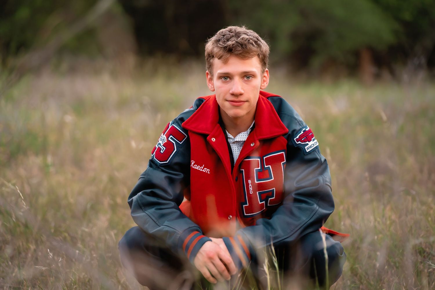 Teen boy in red varsity jacket crouches in tall grass outdoors.
