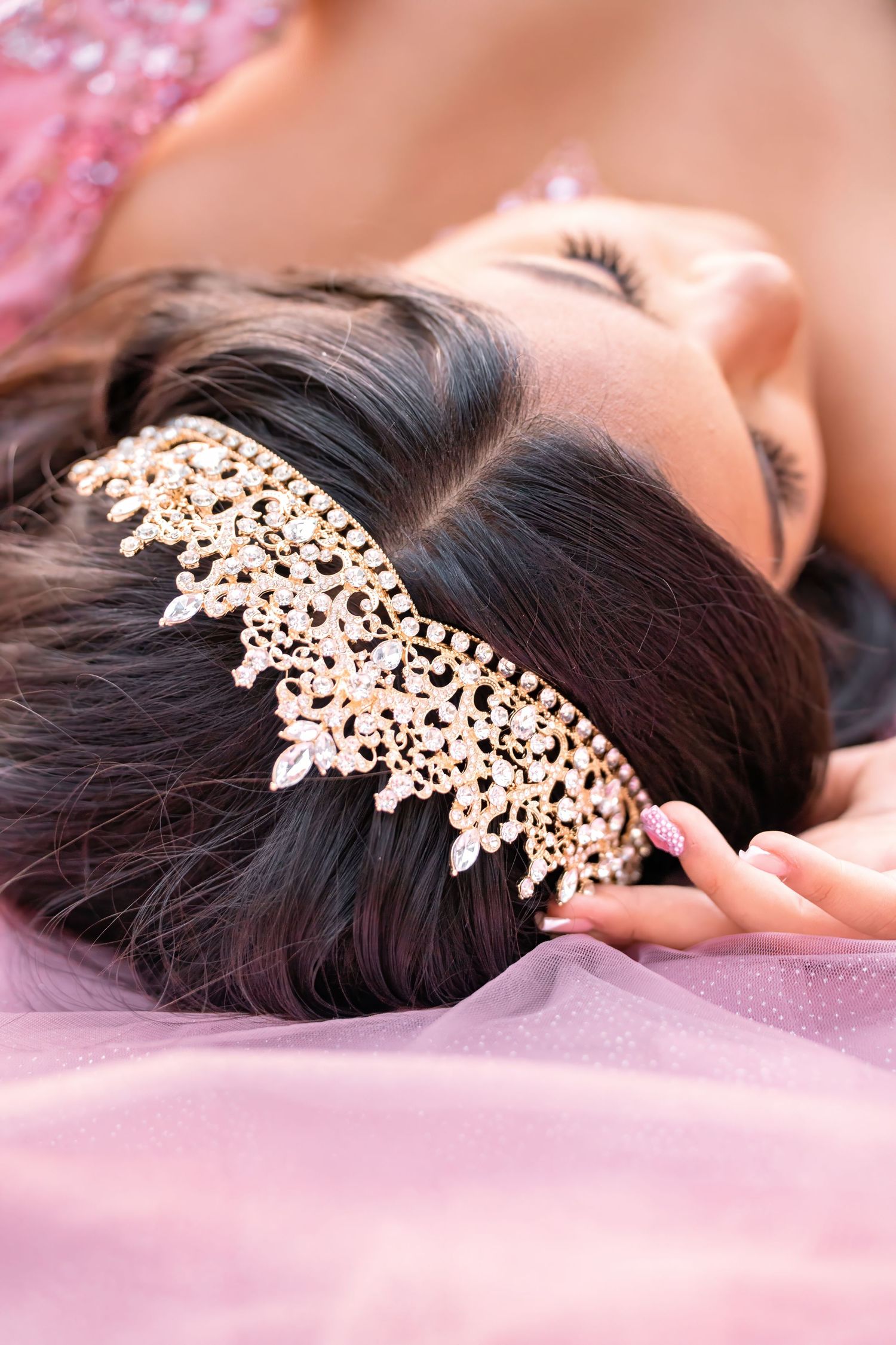 Close-up of ornate gold crystal tiara headpiece in dark hair on pink tulle fabric.