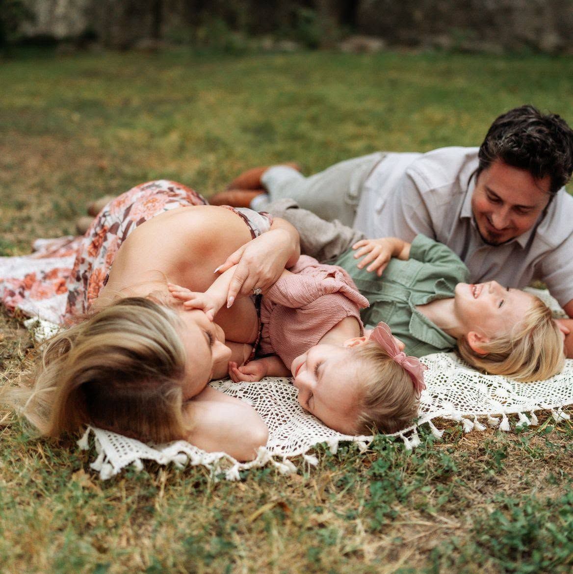 A happy family of four lying on a blanket in the grass, laughing and playing together outdoors.