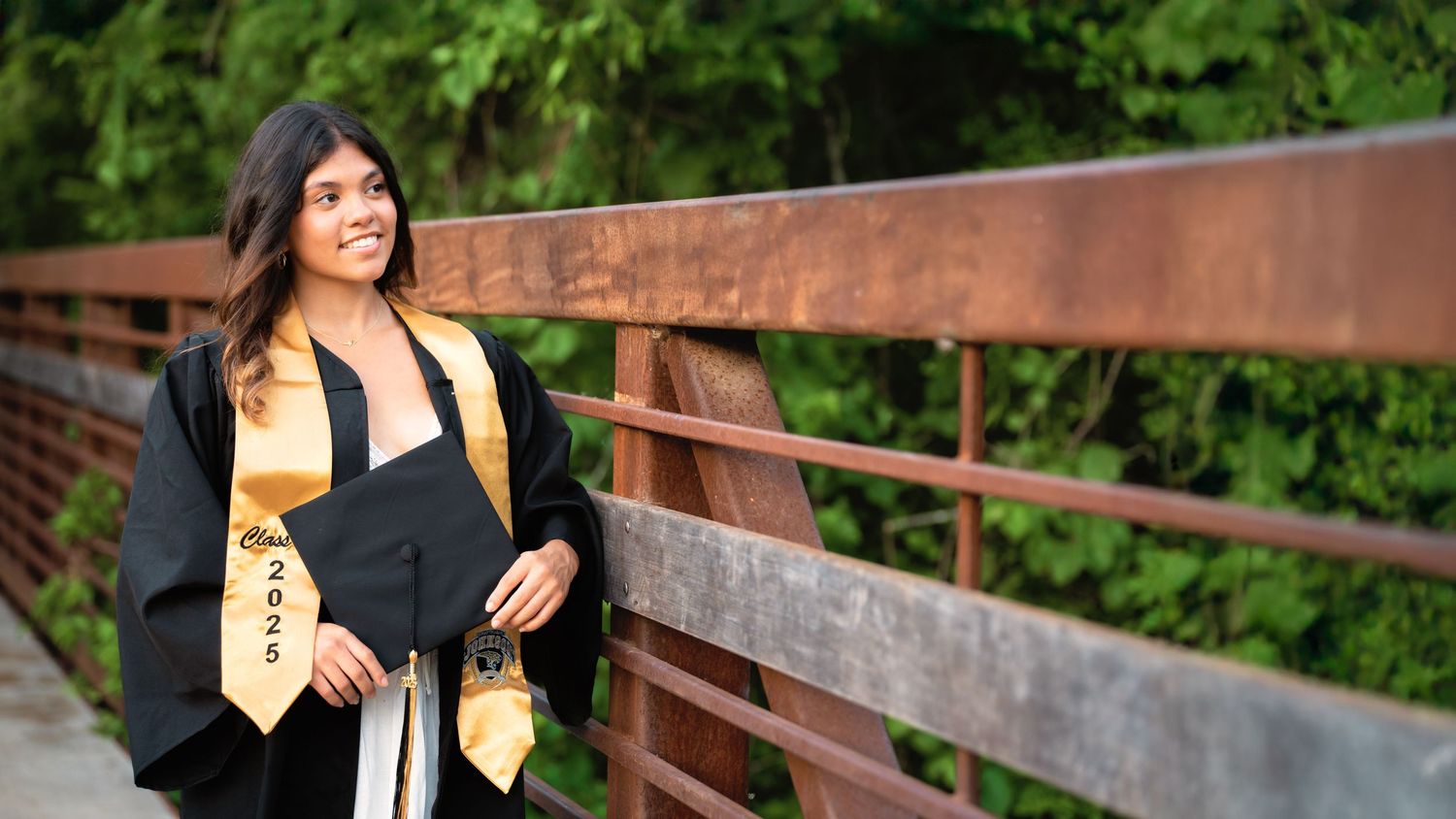 Young woman in black graduation cap and gown with gold sash smiling on a bridge in San Marcos surrounded by greenery.