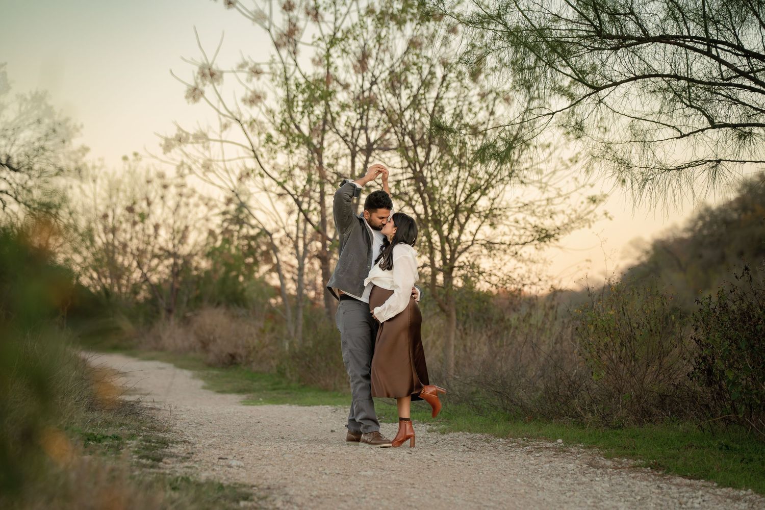 A romantic couple shares a tender moment at sunset on a tree-lined dirt path during a golden hour photoshoot.