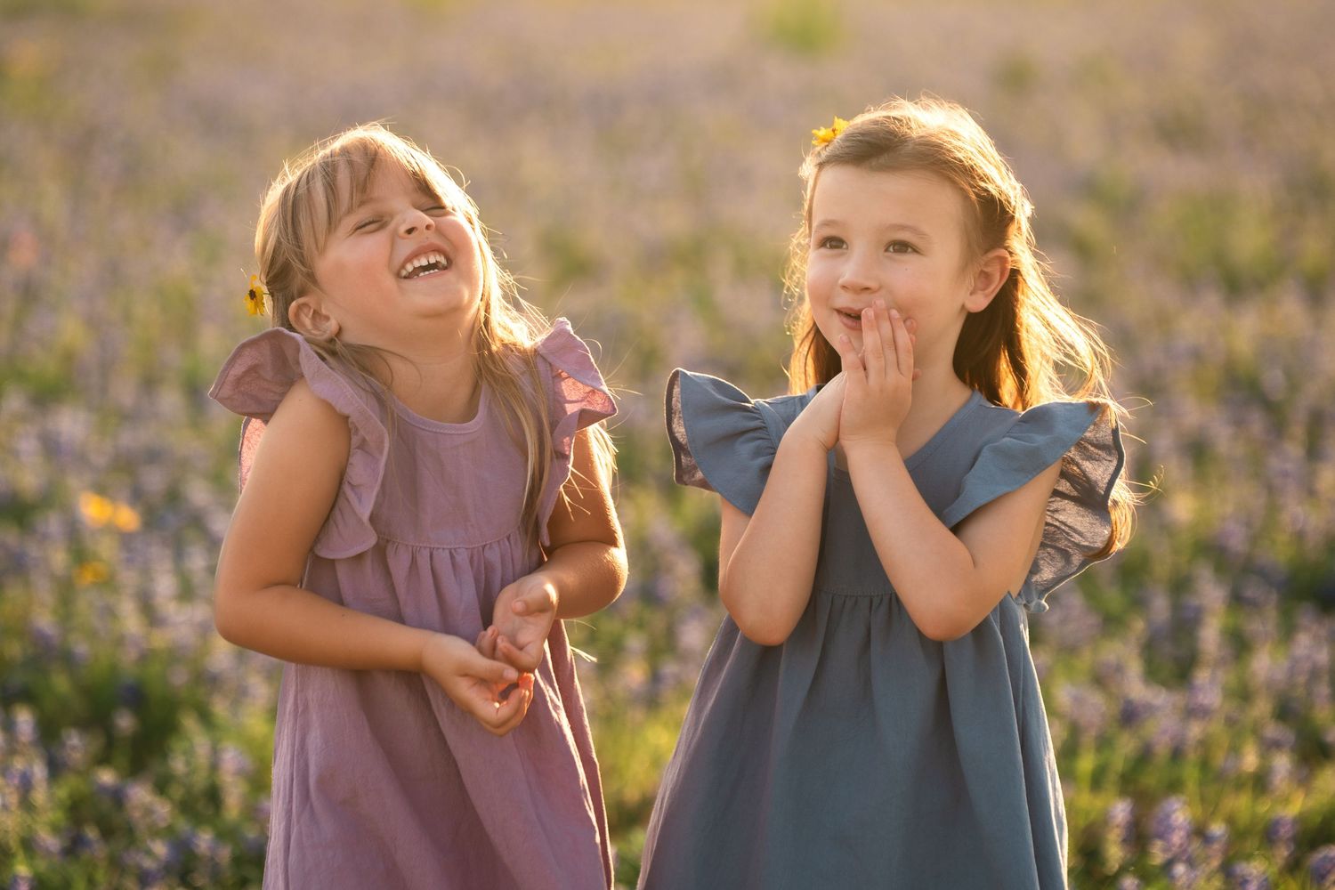 Two children in casual clothing laugh and play together in a sunlit field with purple wildflowers.