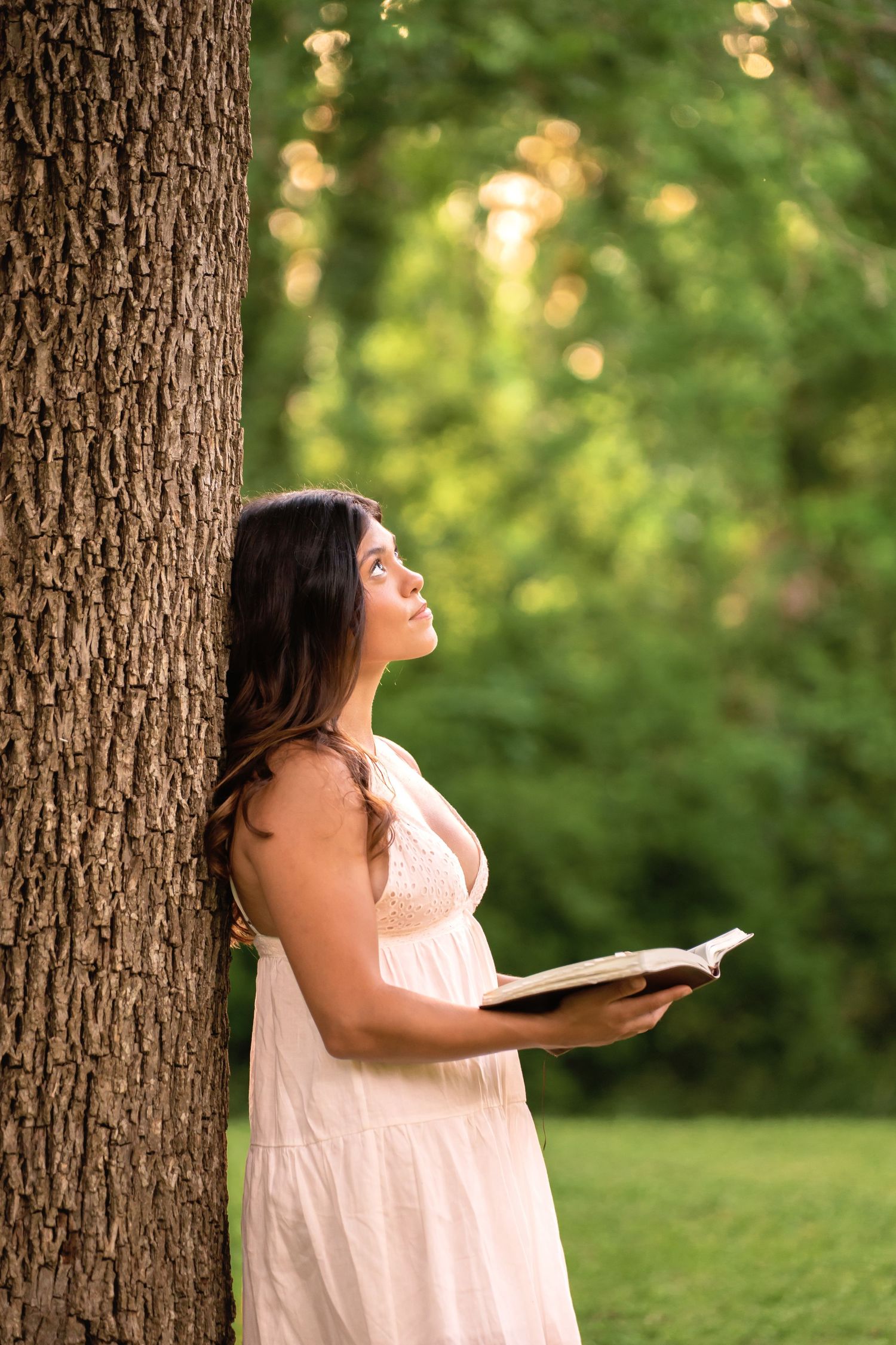 Young woman in white dress leaning against tree, holding open book, looking up in serene outdoor park setting.