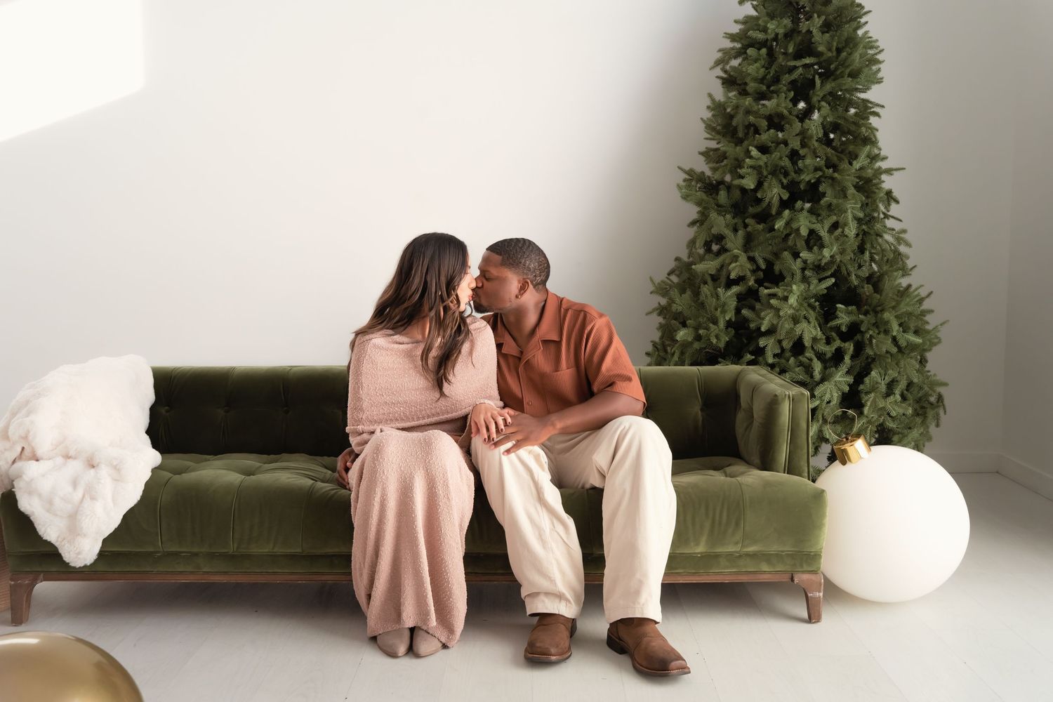 Couple sharing a romantic moment on a green velvet sofa near a Christmas tree in a bright white studio.