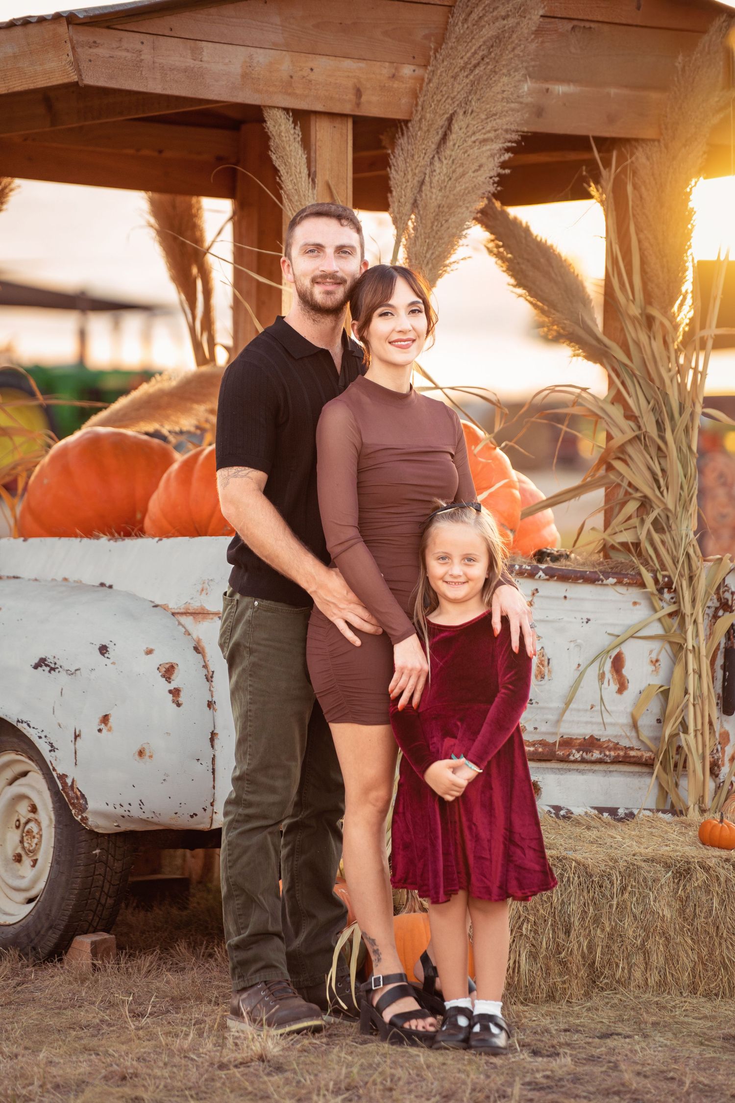 Family of three posing at a pumpkin patch in autumn, parents and young girl in fall attire at golden hour.