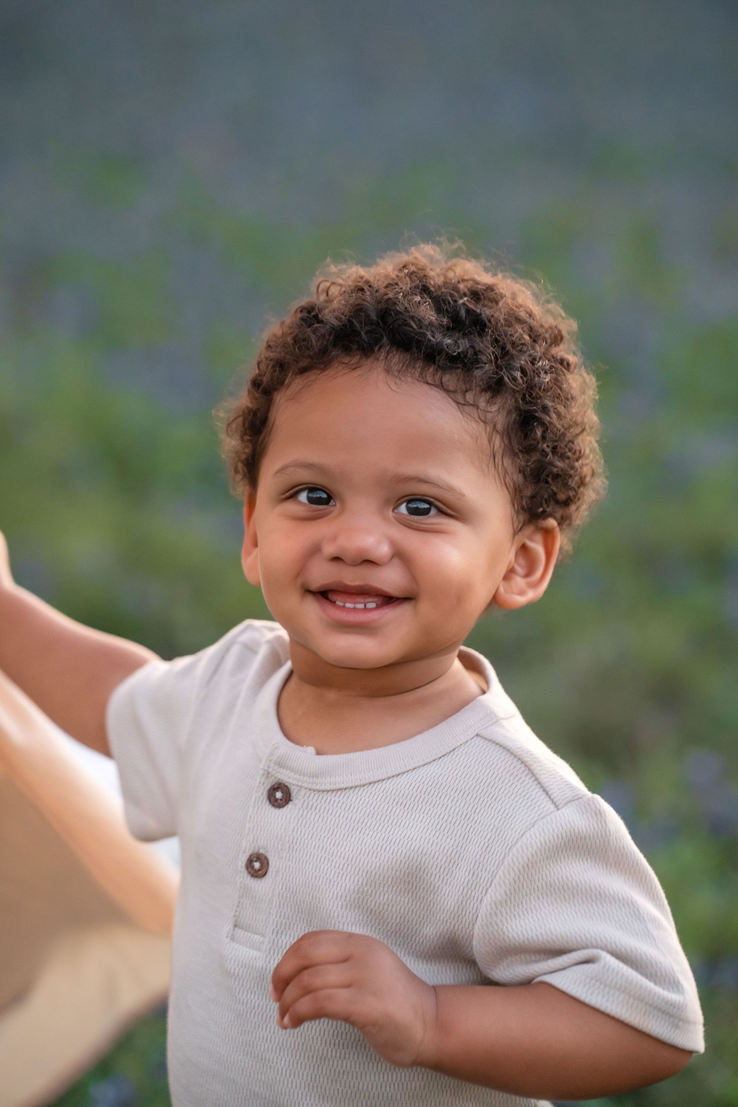 A happy toddler wearing a cream henley shirt smiles brightly outdoors against a blurred natural background.