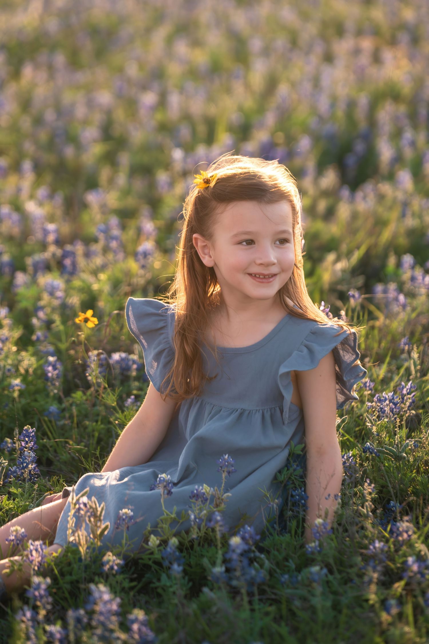 Child wearing blue dress sits among blooming wildflowers in a sunlit meadow during golden hour.