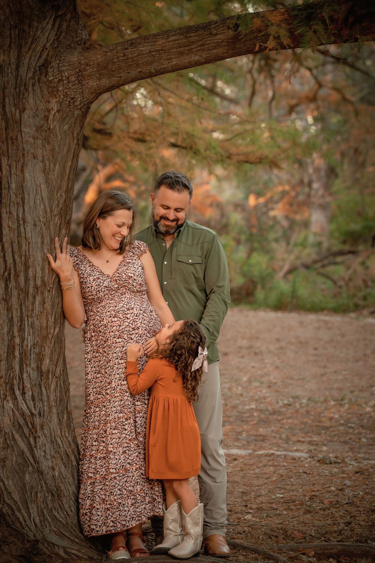 Pregnant woman leans against tree with husband and young daughter kissing her baby bump during outdoor maternity photo session.