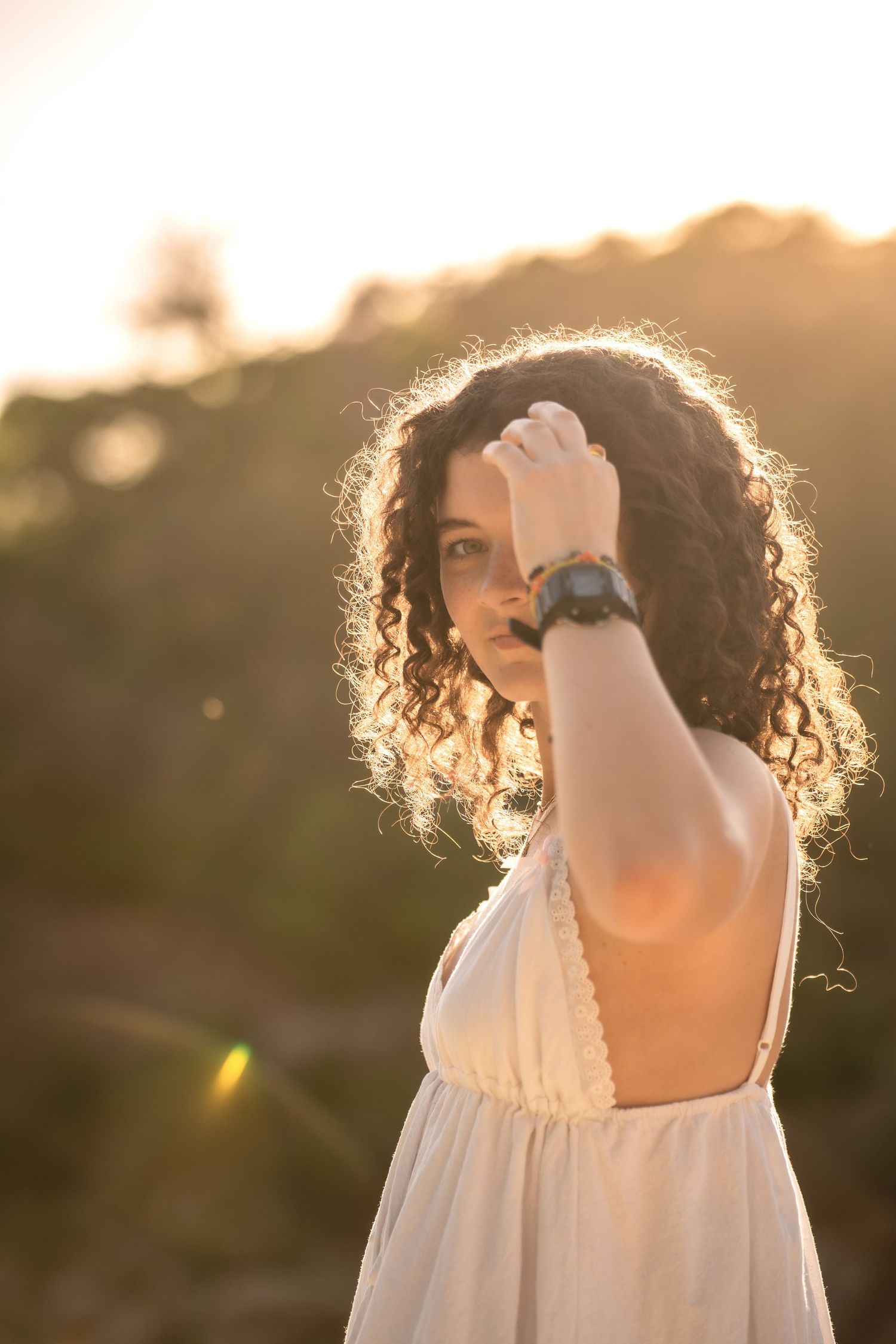 Young woman with curly hair in white sundress shielding eyes from golden sunset light outdoors.