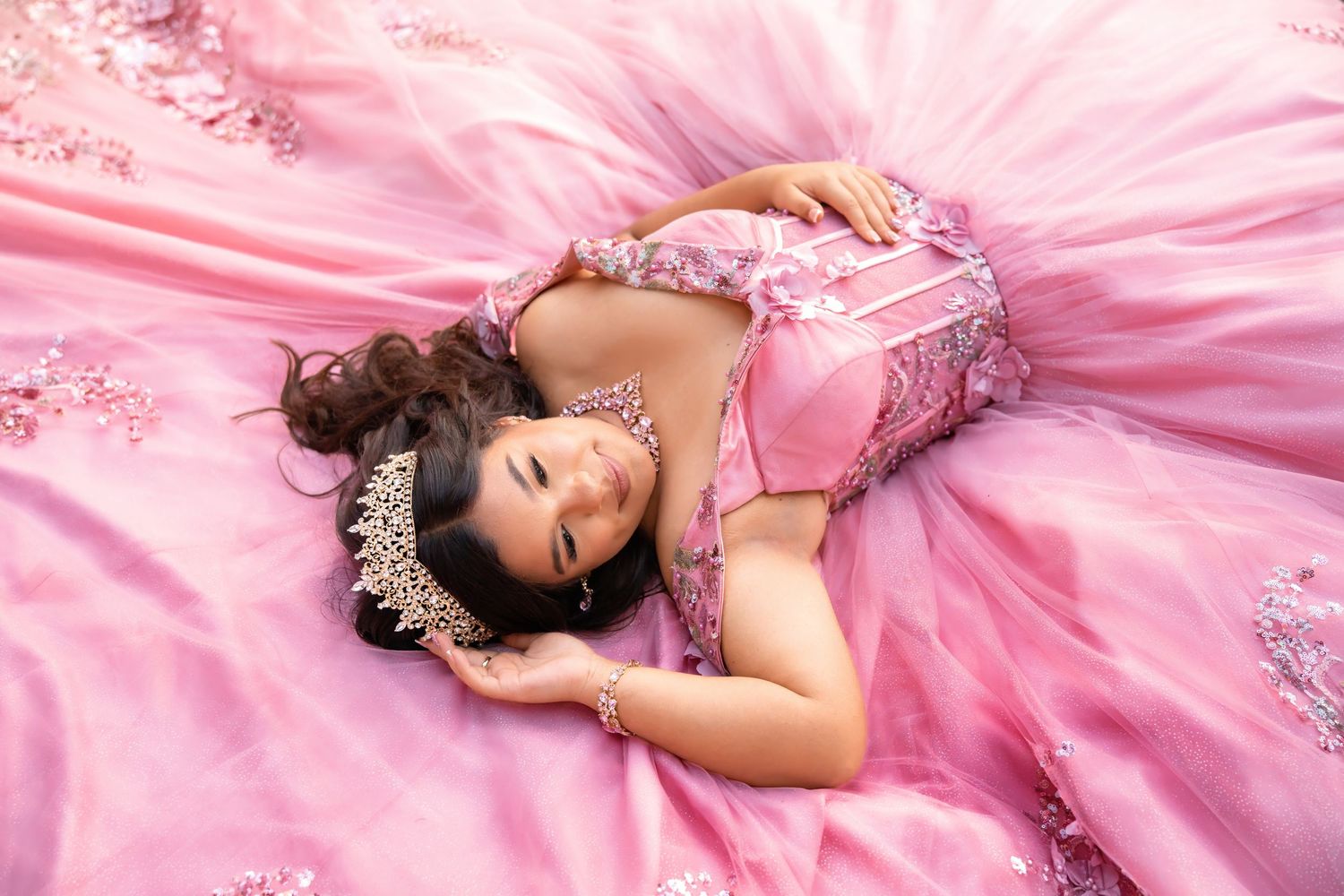 Teen girl in pink quinceañera dress and tiara lies gracefully on billowing pink tulle fabric.