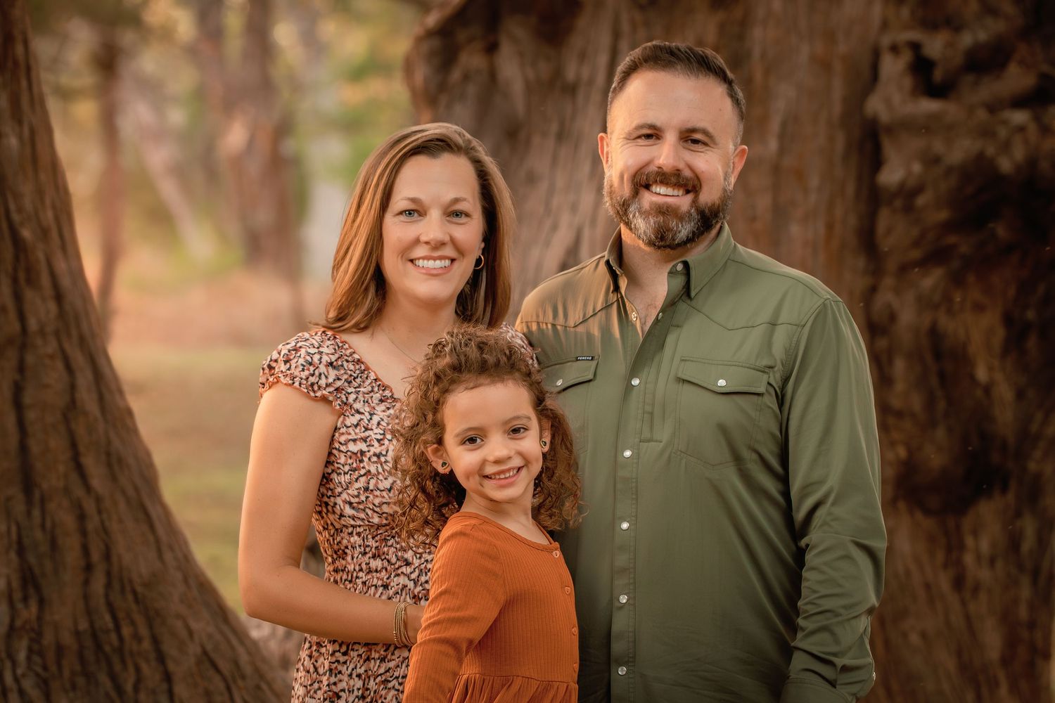 Family of three smiling outdoors near a large tree, parents and young child posing for a portrait photo.