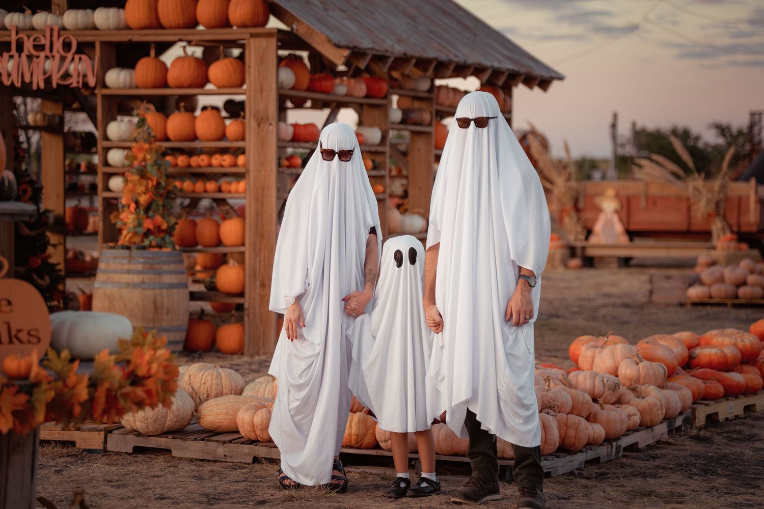 Three people in white ghost costumes with sunglasses pose at a pumpkin patch during Halloween.