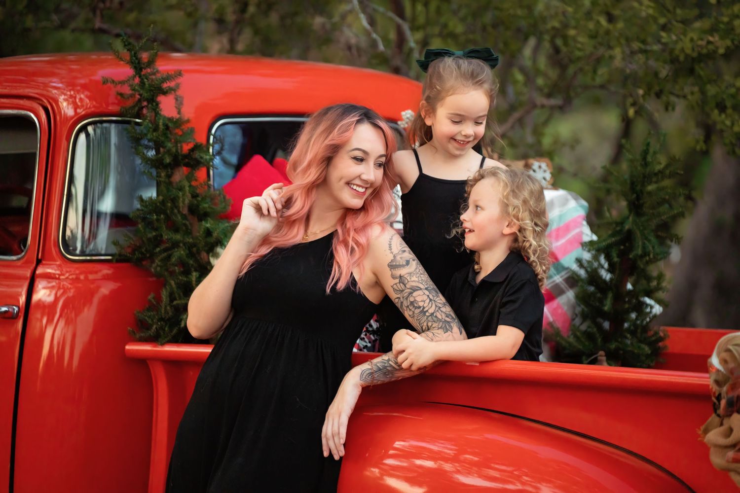 Woman with pink hair and tattoos laughs with two girls leaning on a vintage red truck outdoors.