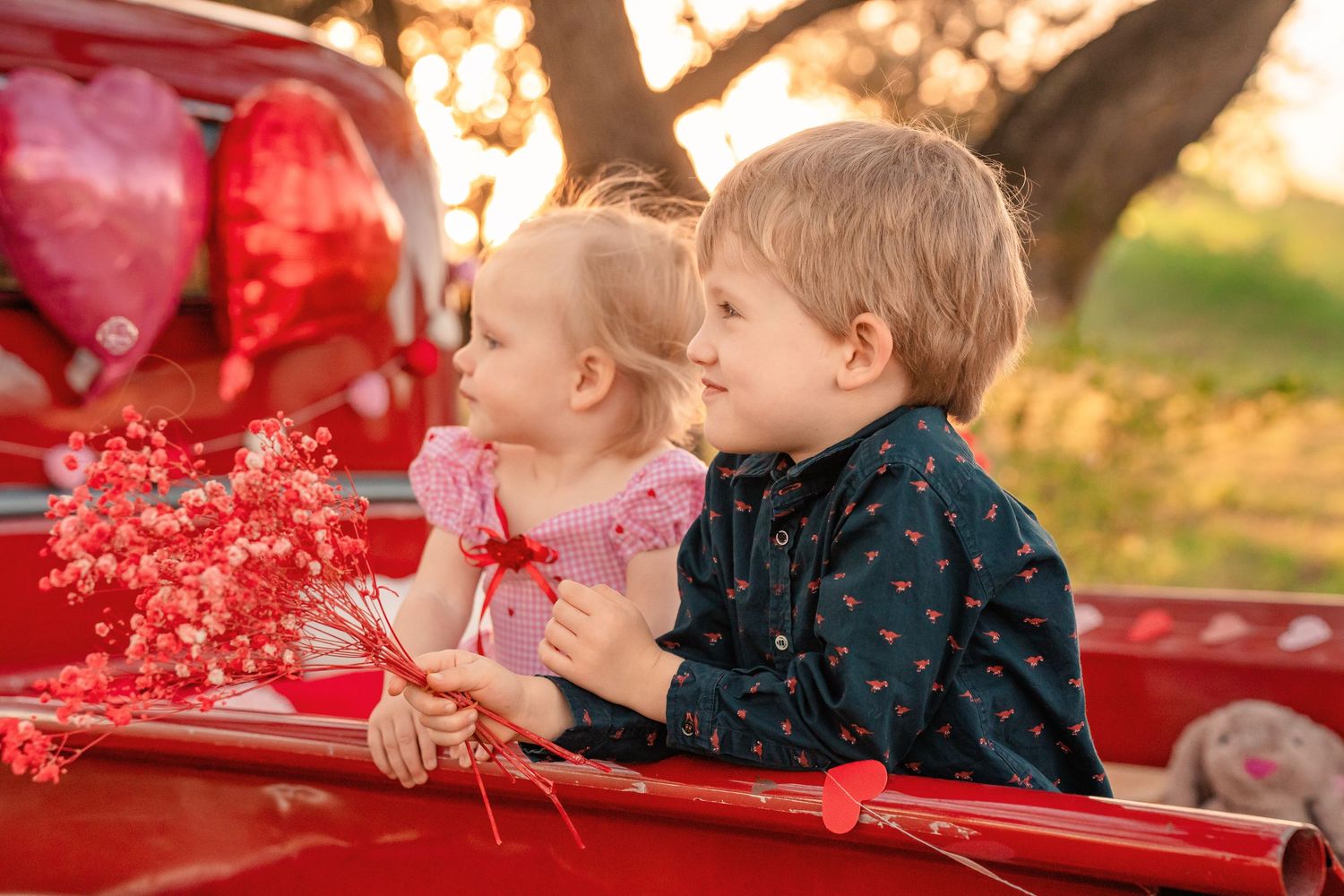 Toddler girl and young boy sit together in a red vintage truck bed holding pink baby's breath flowers with heart balloons at sunset.