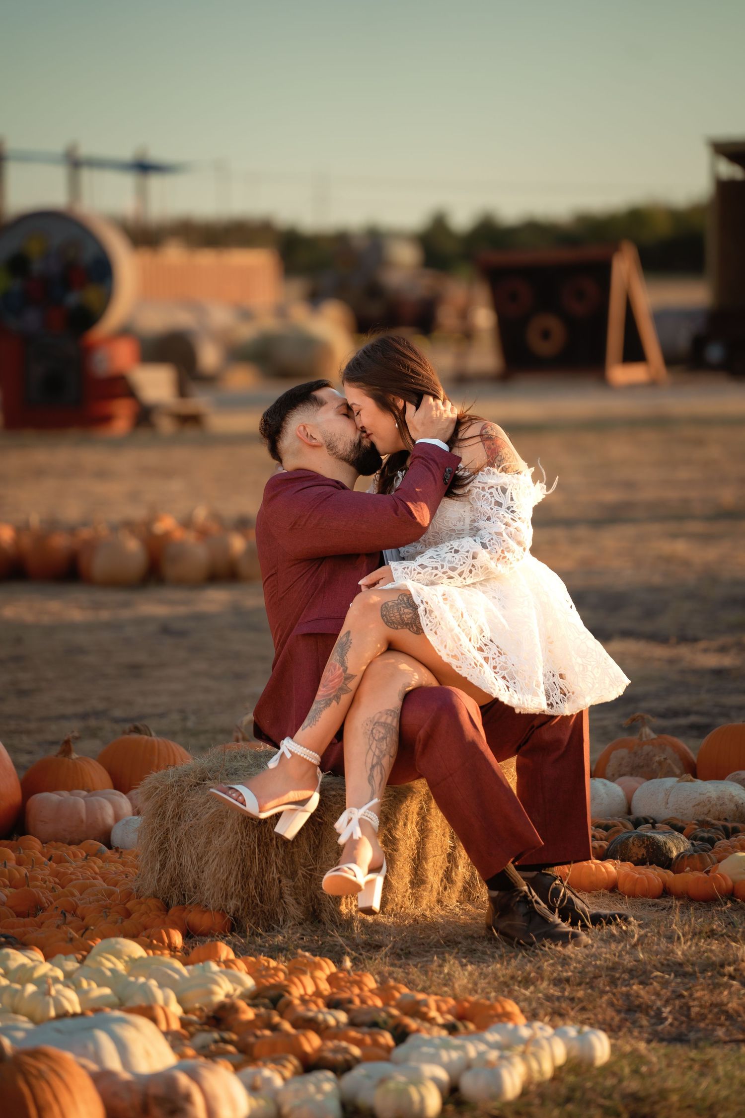 Romantic couple sitting on hay bale at pumpkin patch, sharing a kiss in warm autumn sunset light.