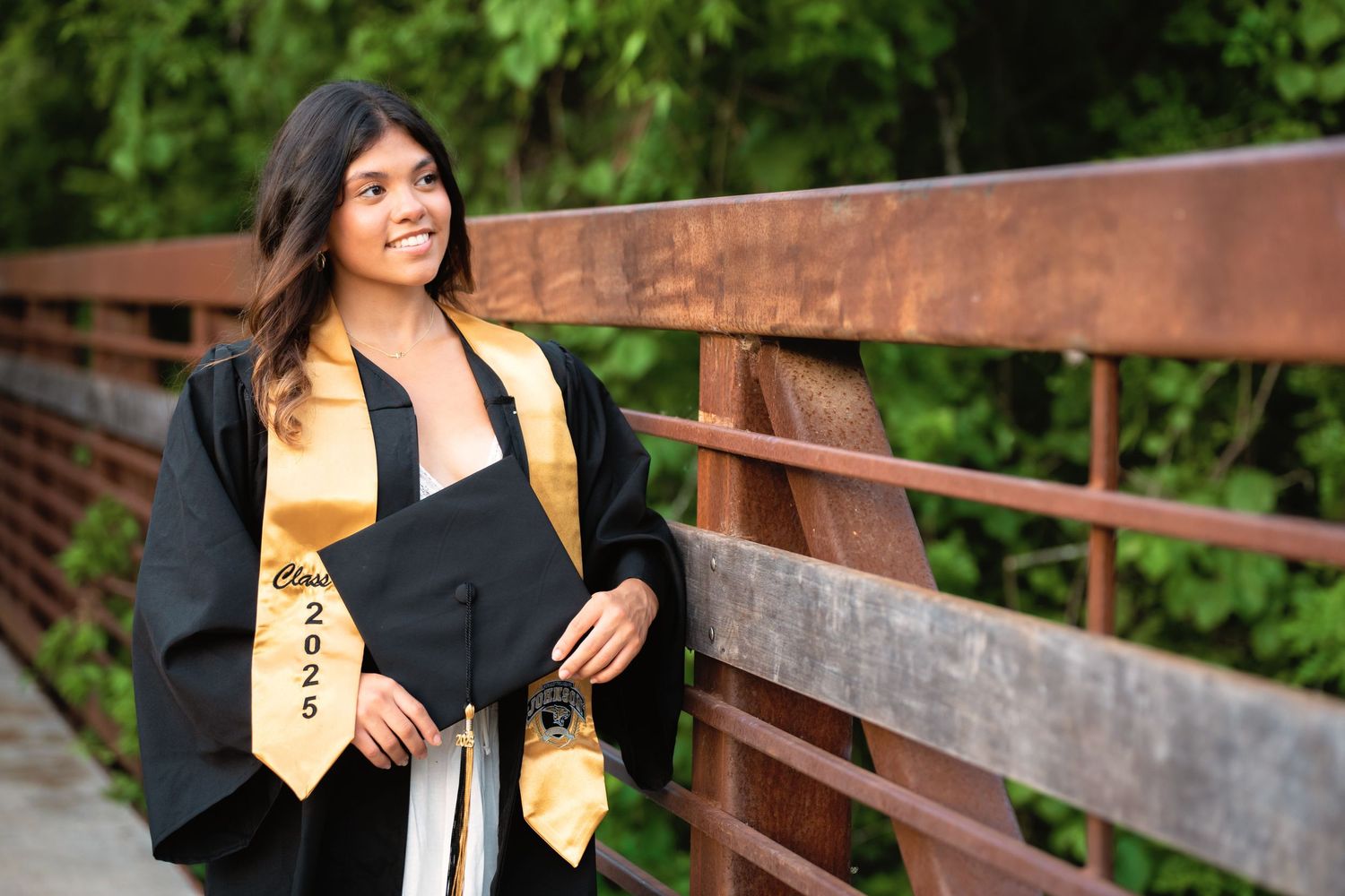A smiling young woman in black graduation gown and gold stole poses on a rustic bridge surrounded by lush greenery.